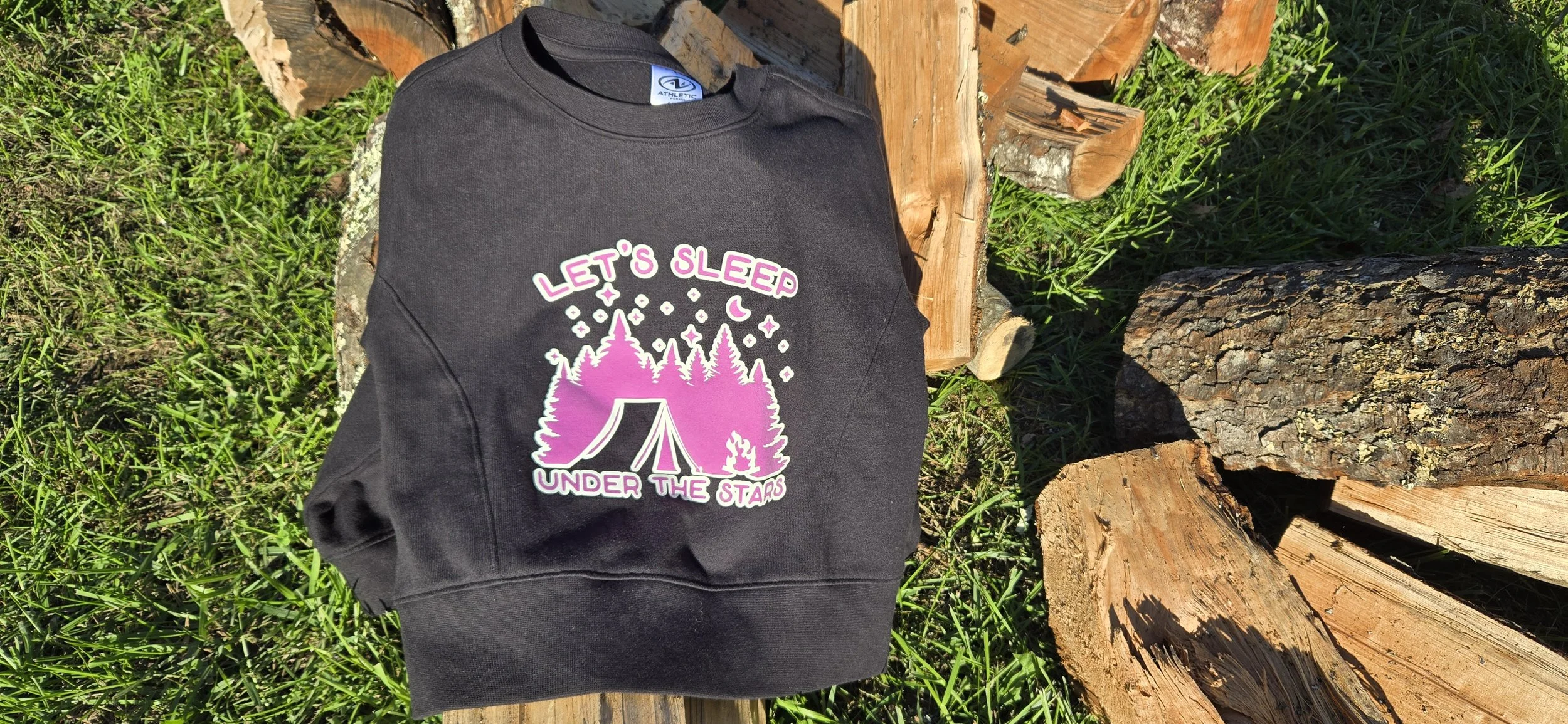 Black sweatshirt with a pink Let's Sleep Under the Stars design of a tent and pine trees, laid on grass beside firewood