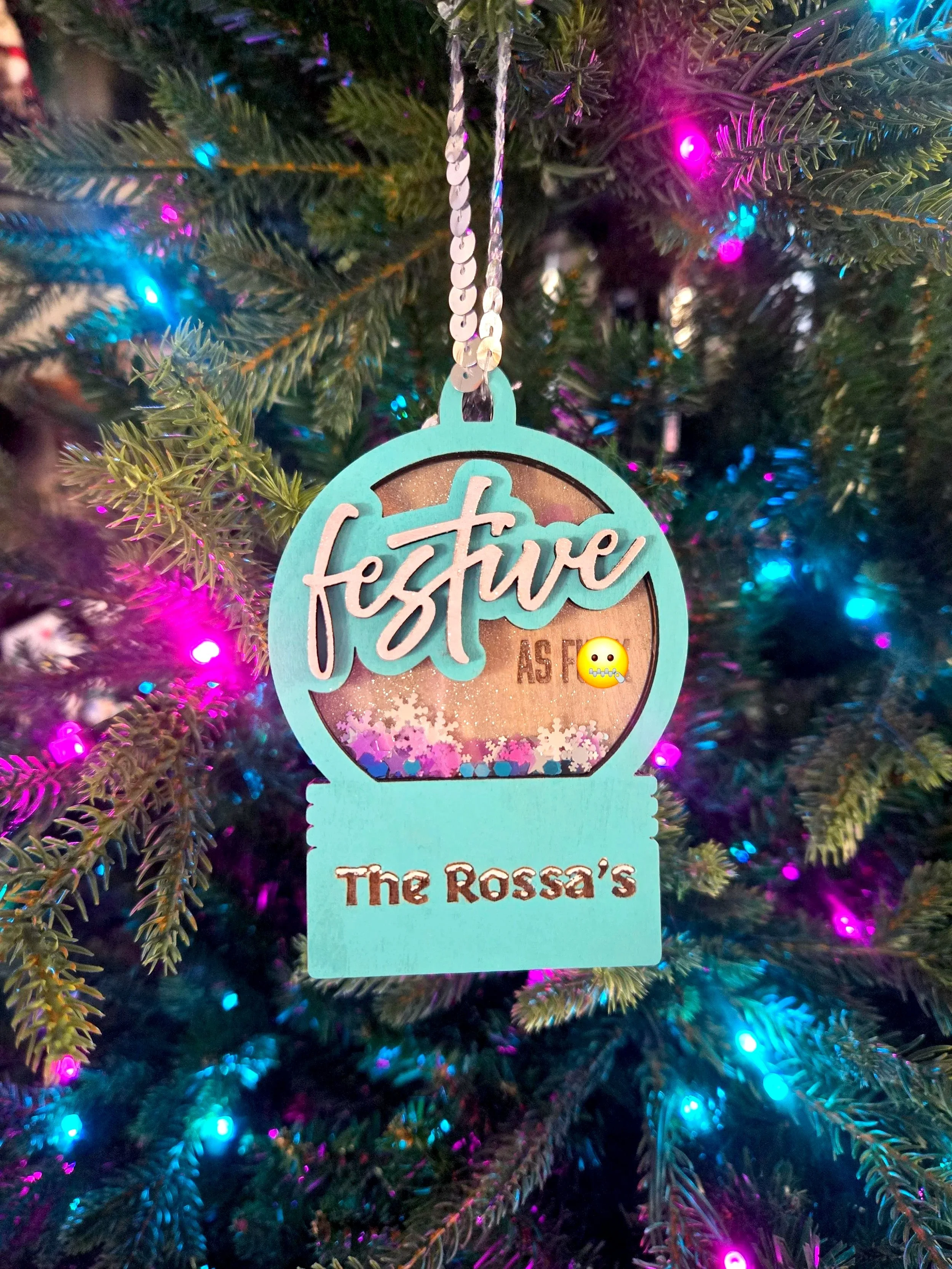 Teal shaker ornament reading Festive AF with a family name below, filled with snowflake confetti, hanging on a lit Christmas tree