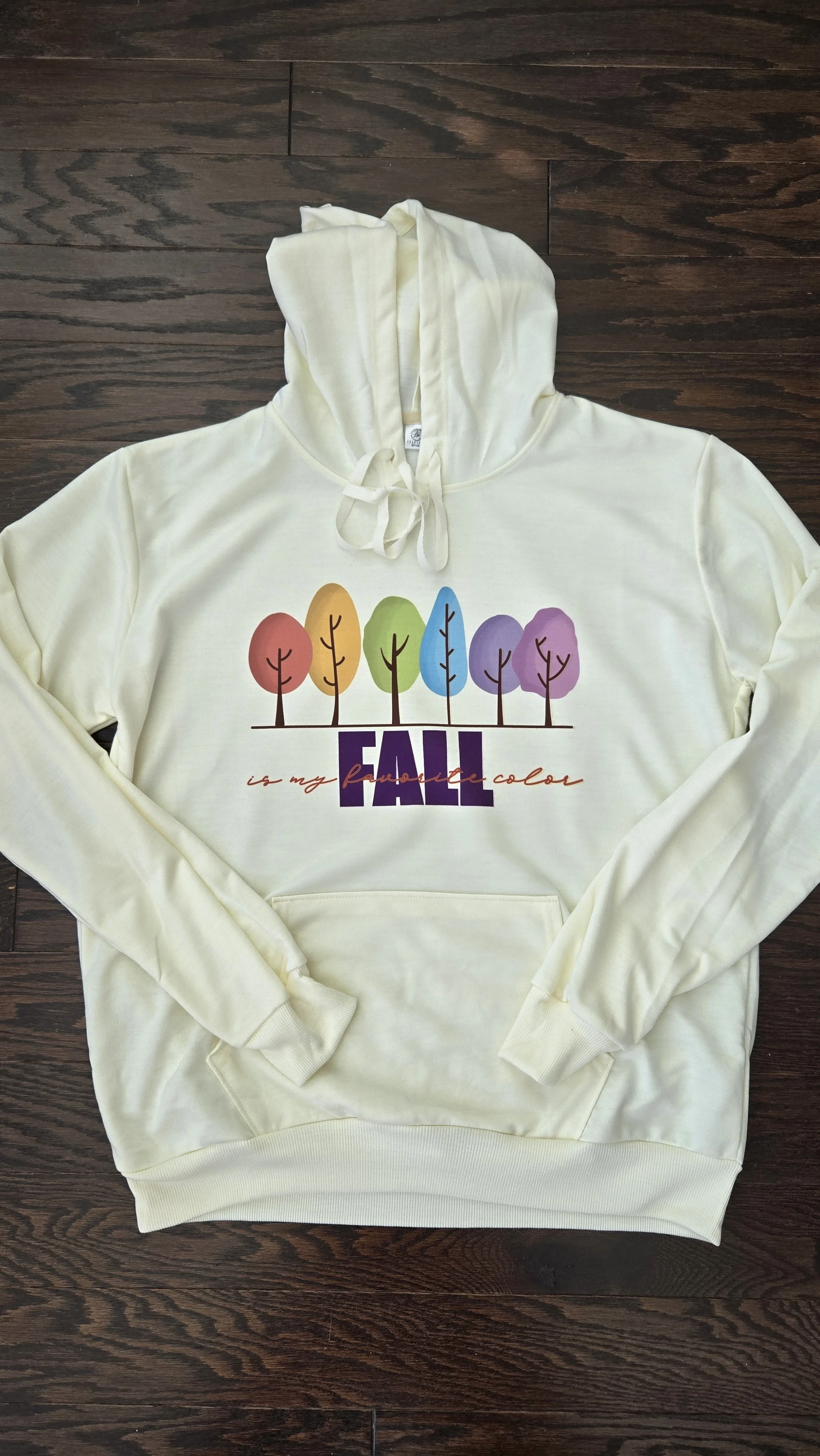 Cream hoodie printed with a row of rainbow watercolor trees and the word Fall in purple