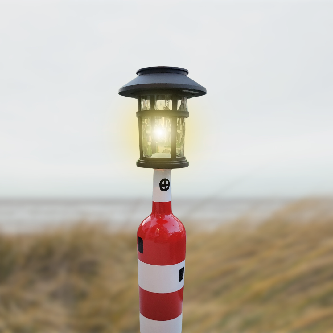 Red and white striped wine bottle lighthouse lamp topped with a glowing solar lantern, set against beach dunes