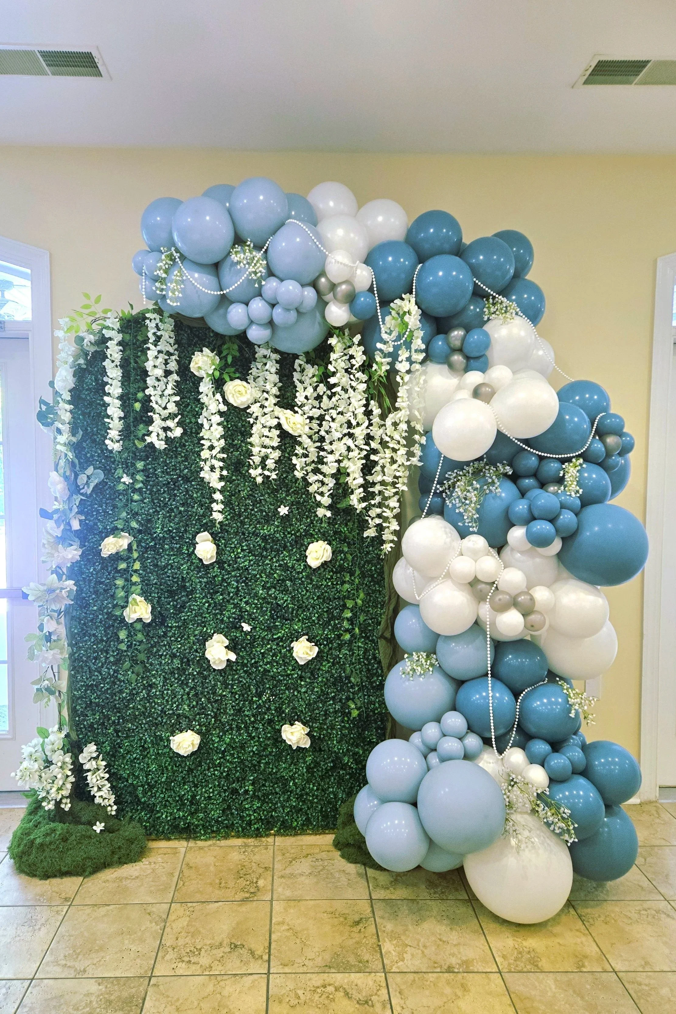 A decorative floral arrangement with a wall of green foliage and white roses, decorated with white floral garlands, on the left side, and a large arch of blue, white, and silver balloons with small flowers and pearl strings on the right side, placed indoors on tiled flooring.