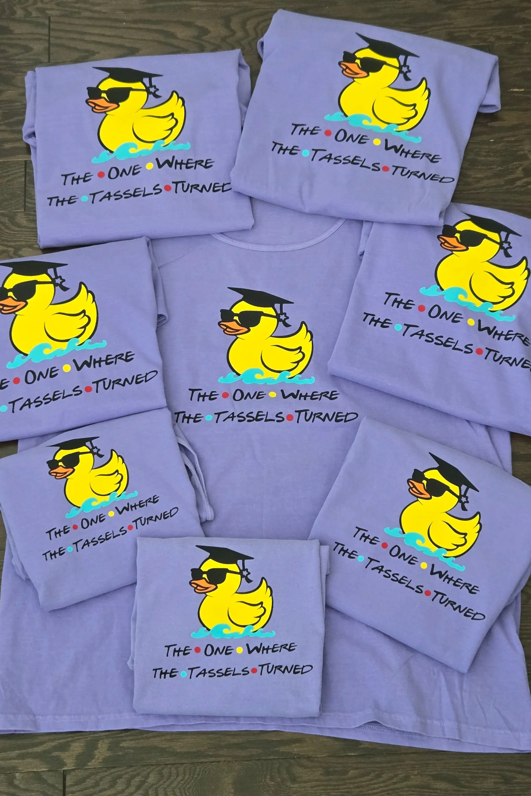 Multiple purple T-shirts featuring a cartoon rubber duck wearing sunglasses and a graduation cap. The text on the shirts reads, 'The one where the tassels turned' in black with colorful dots.