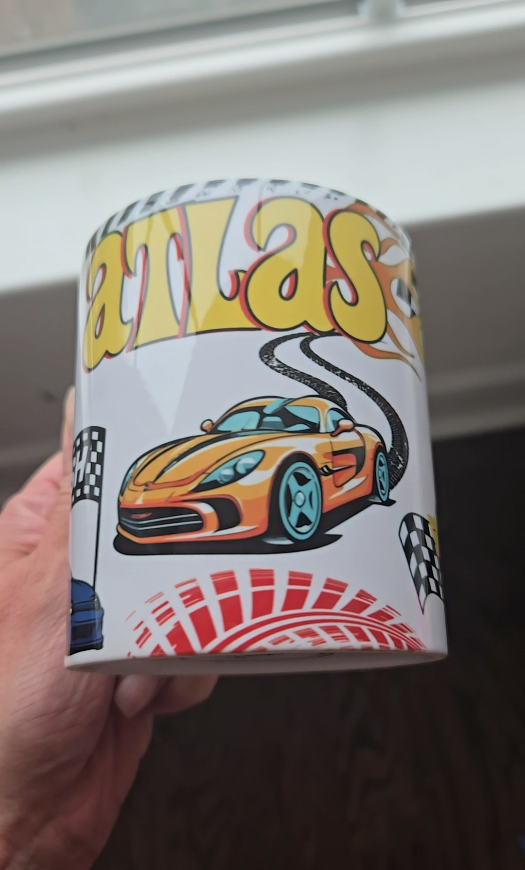Hand holding a white mug printed with a child's name above an orange sports car, a racetrack, and checkered flags