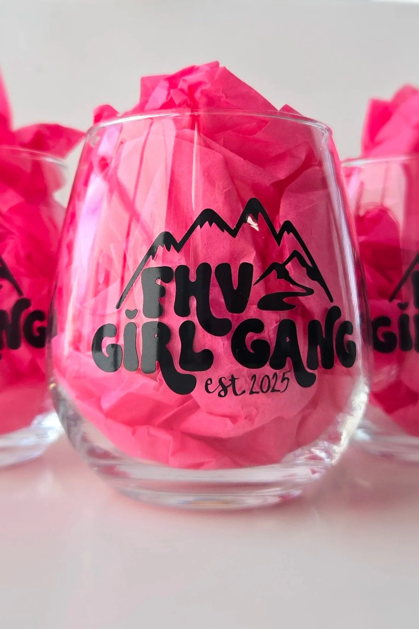 Close-up of a clear glass cup with pink tissue paper inside, decorated with a black mountain outline and the words "Girls Gifting" and "est. 2025".