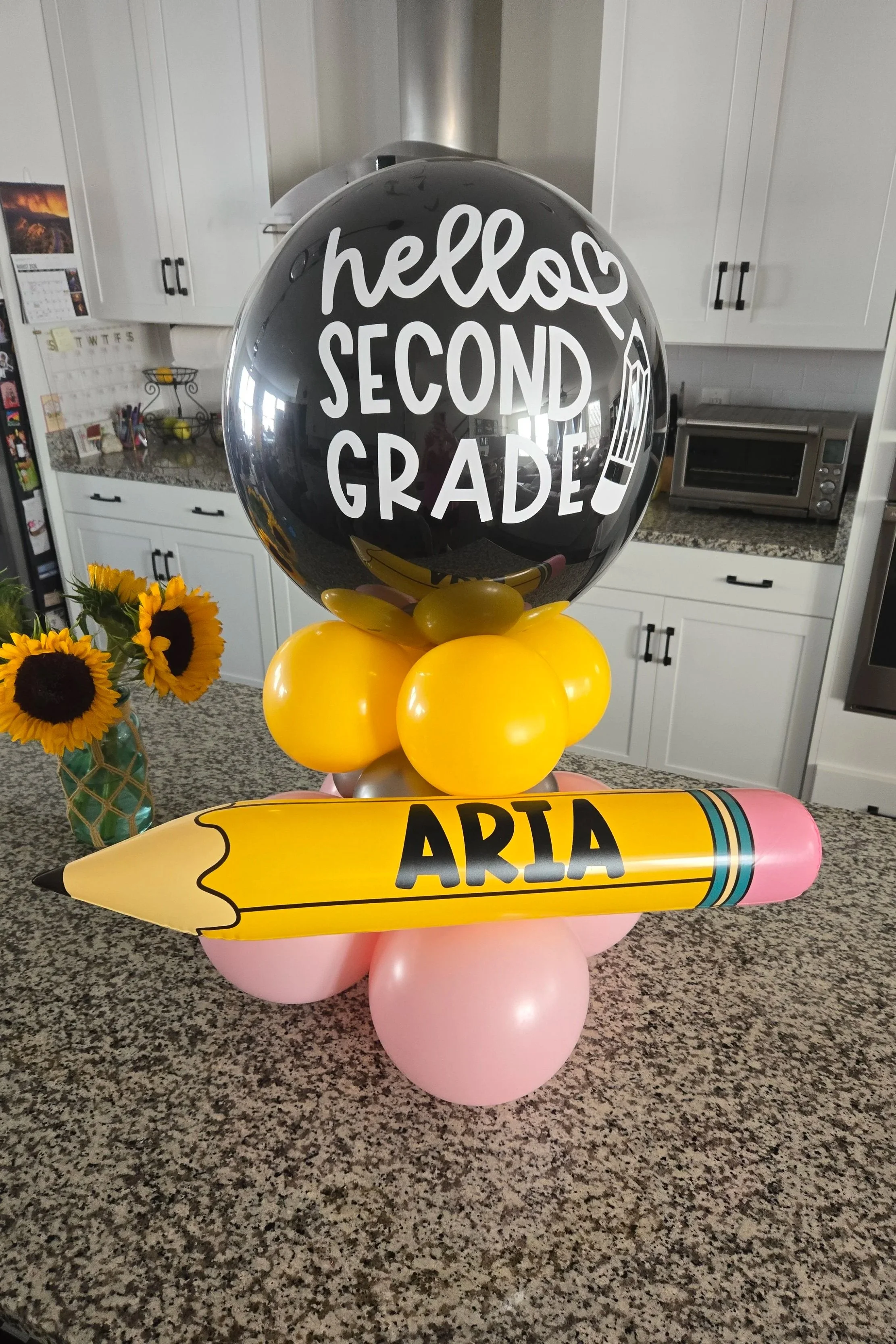 Decorative bouquets with sunflowers, a cluster of yellow balloons, a black balloon with 'hello SECOND GRADE' written on it, a pink balloon, and a large yellow crayon-shaped decoration with the name 'ARIA' written on it, on a kitchen countertop.