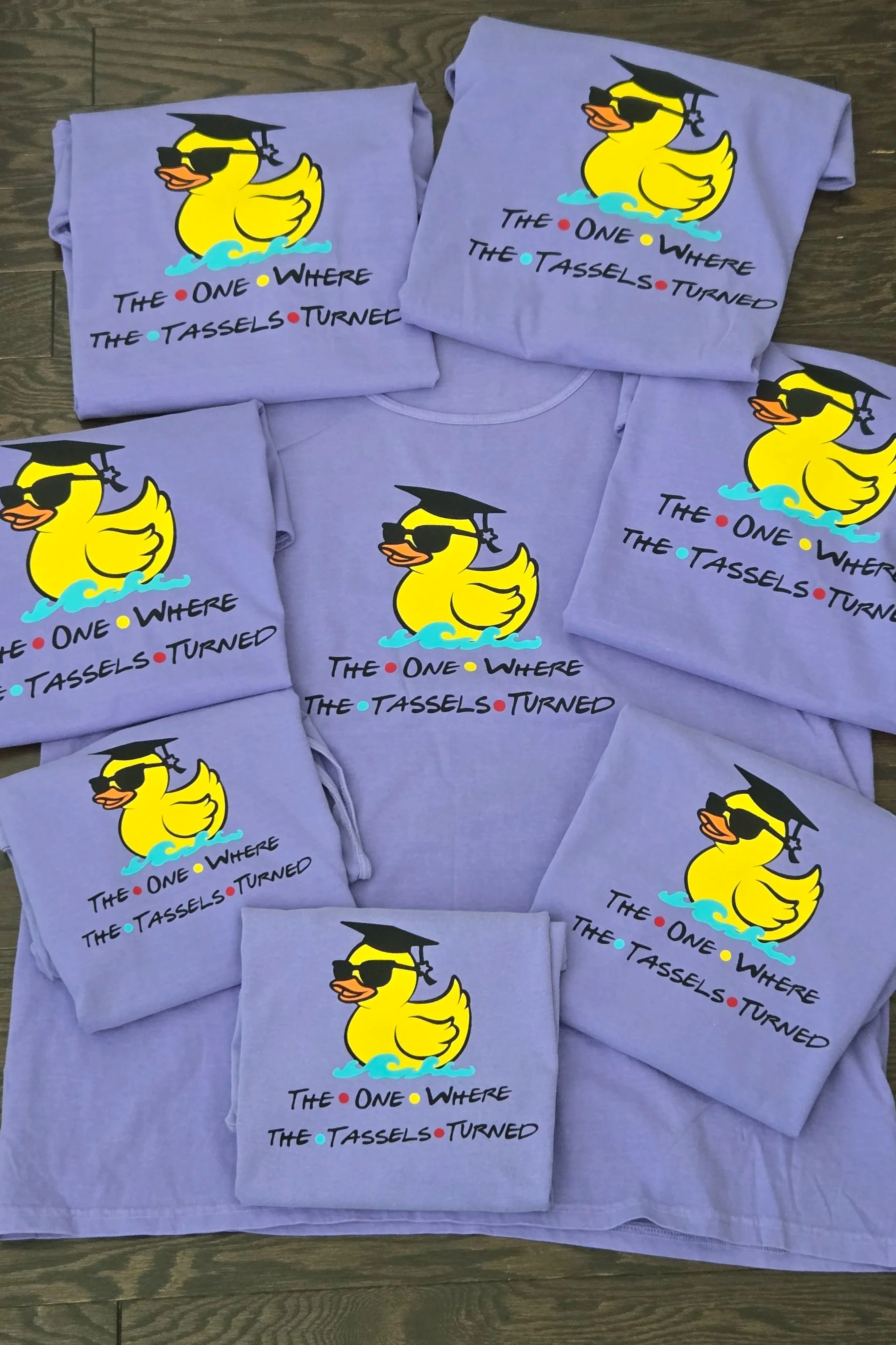Multiple purple T-shirts featuring a cartoon rubber duck wearing sunglasses and a graduation cap. The text on the shirts reads, 'The one where the tassels turned' in black with colorful dots.