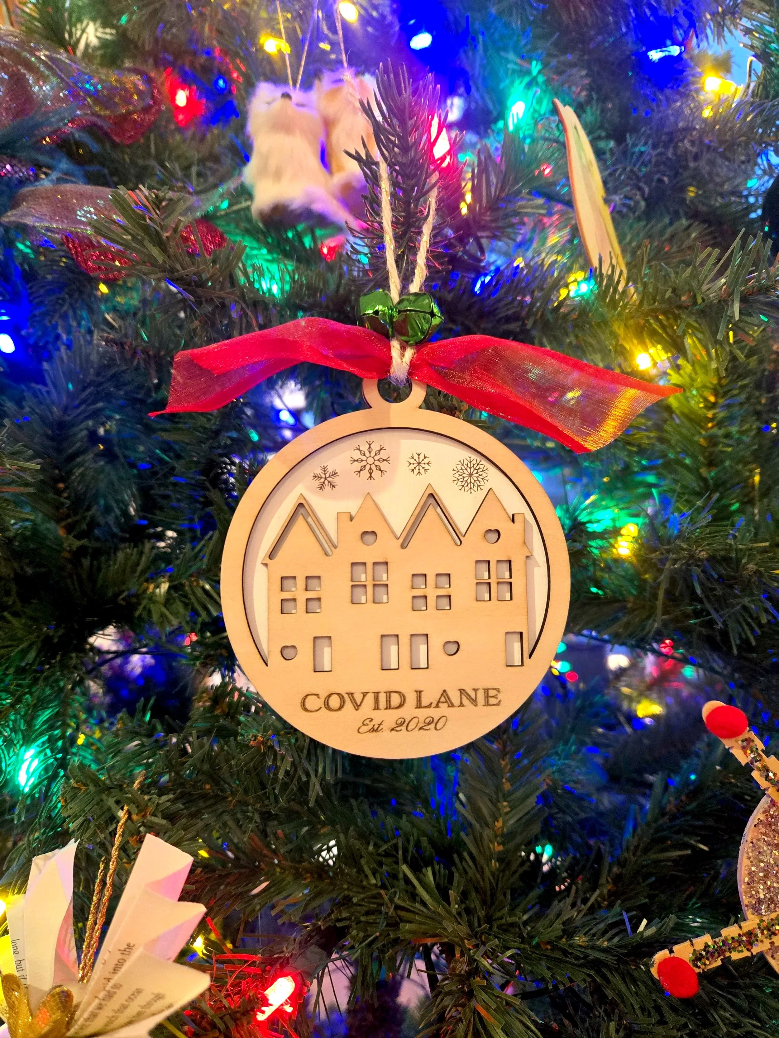 Round wooden ornament engraved with a row of townhomes, a street name, and the year established, hanging on a lit Christmas tree
