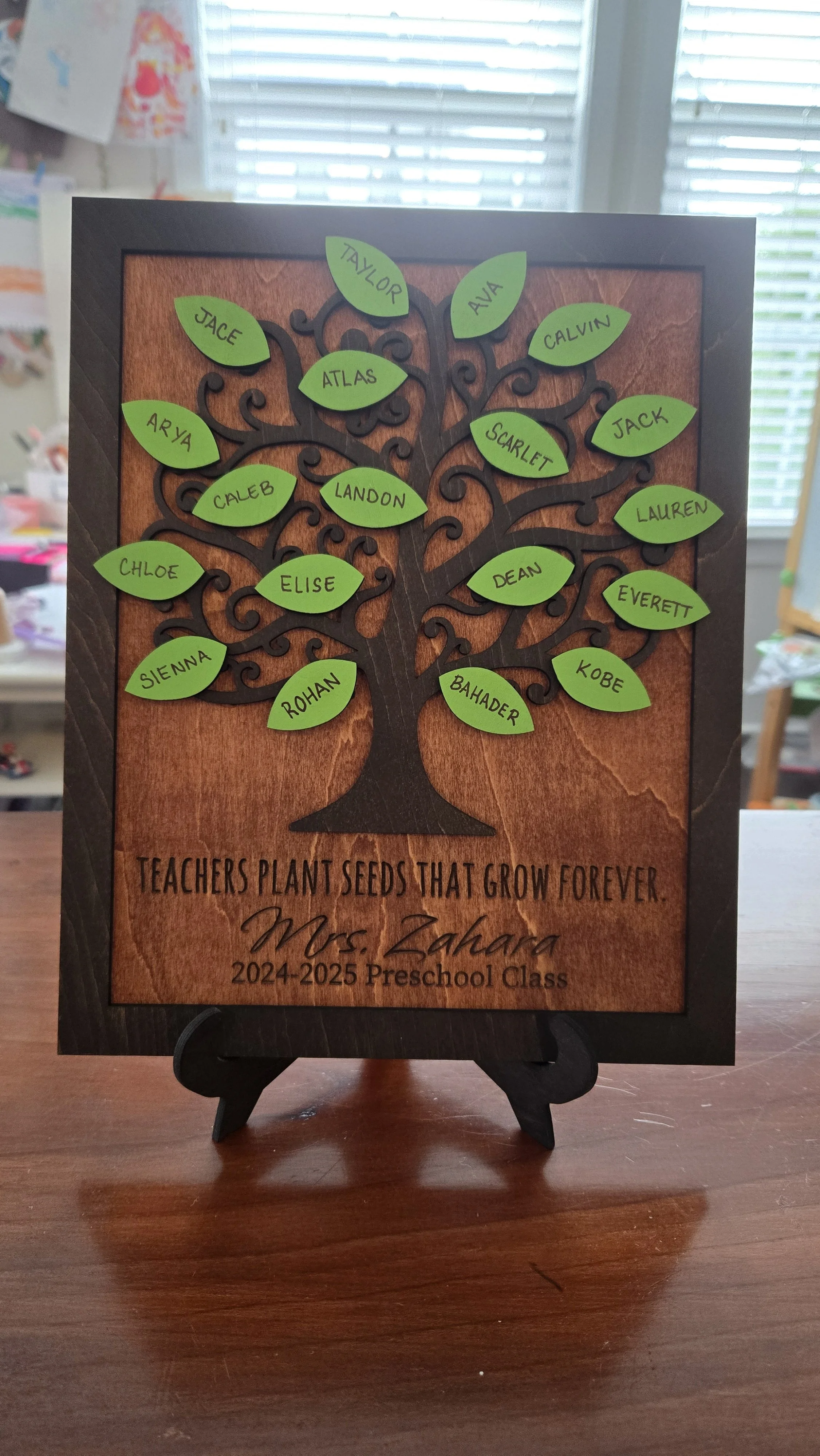 A decorative wooden plaque with a tree design, featuring green paper leaves with handwritten names, and a quote at the bottom that reads 'Teachers plant seeds that grow forever.' It is personalized for Mrs. Zahara, a 2024-2025 preschool class teacher, and displayed on a small easel on a wooden surface.