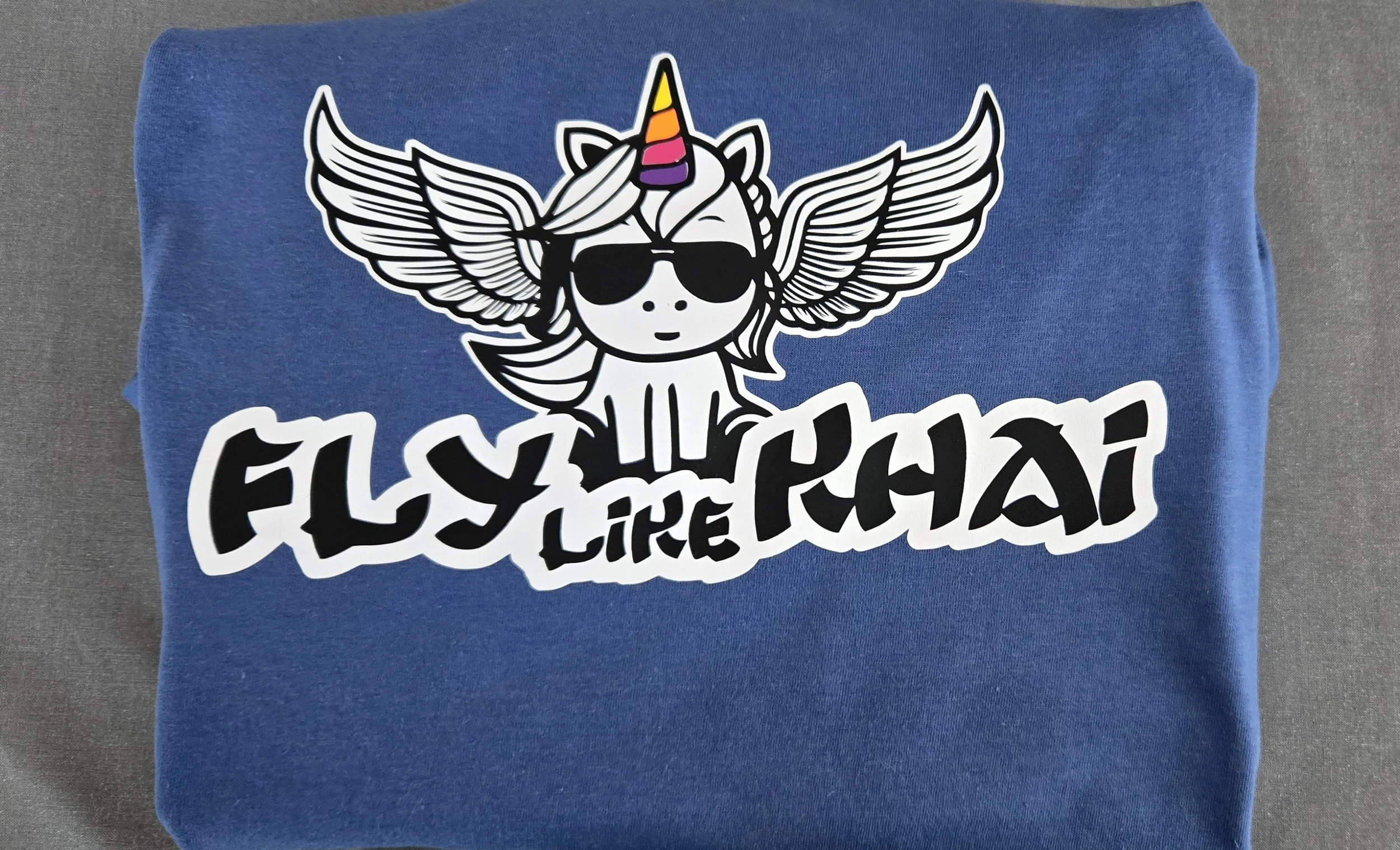 Blue shirt with a winged unicorn wearing sunglasses and the phrase Fly Like Khai
