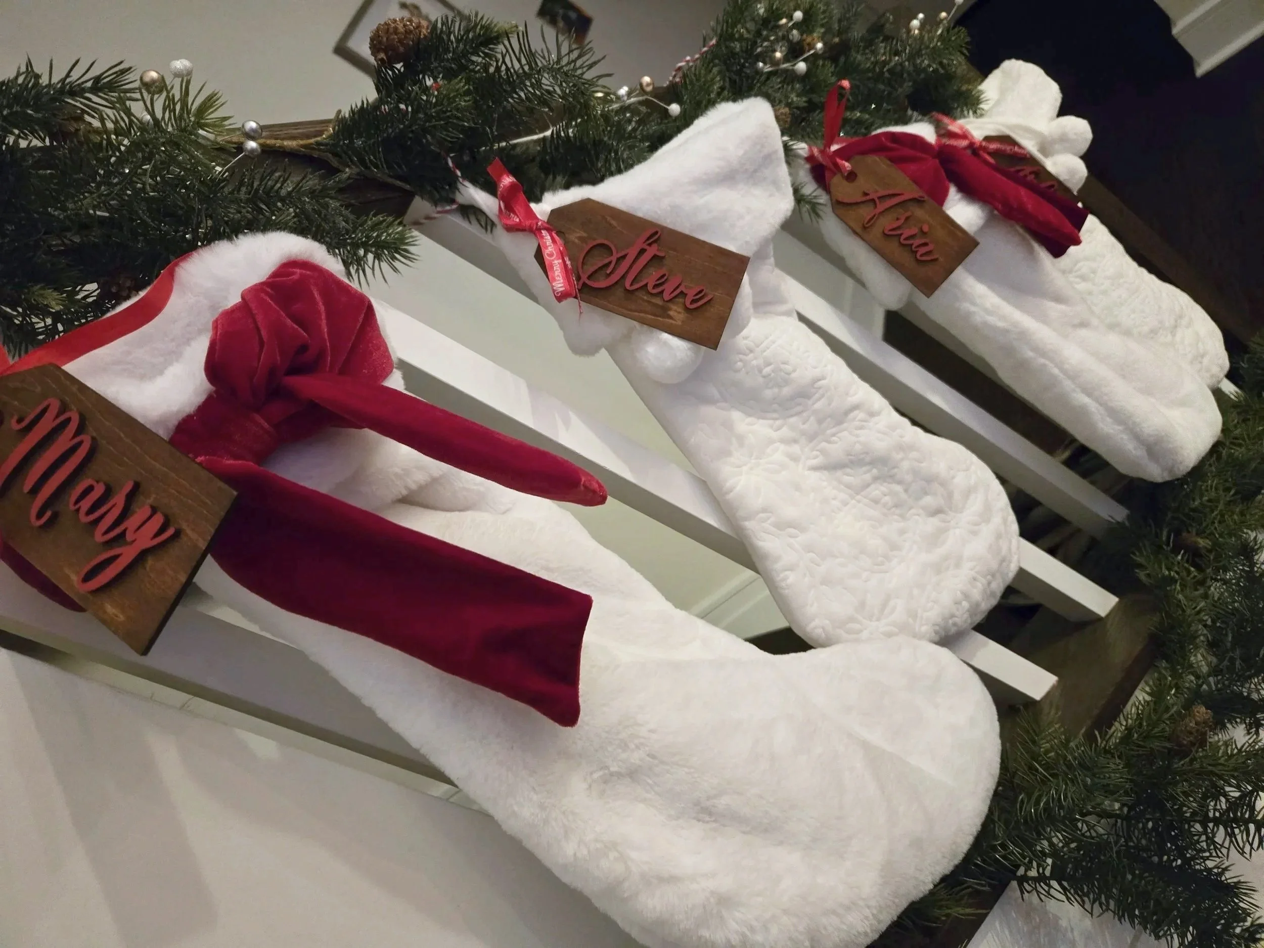 White faux fur Christmas stockings with red velvet bows and wooden name tags in red script, hung along a garland