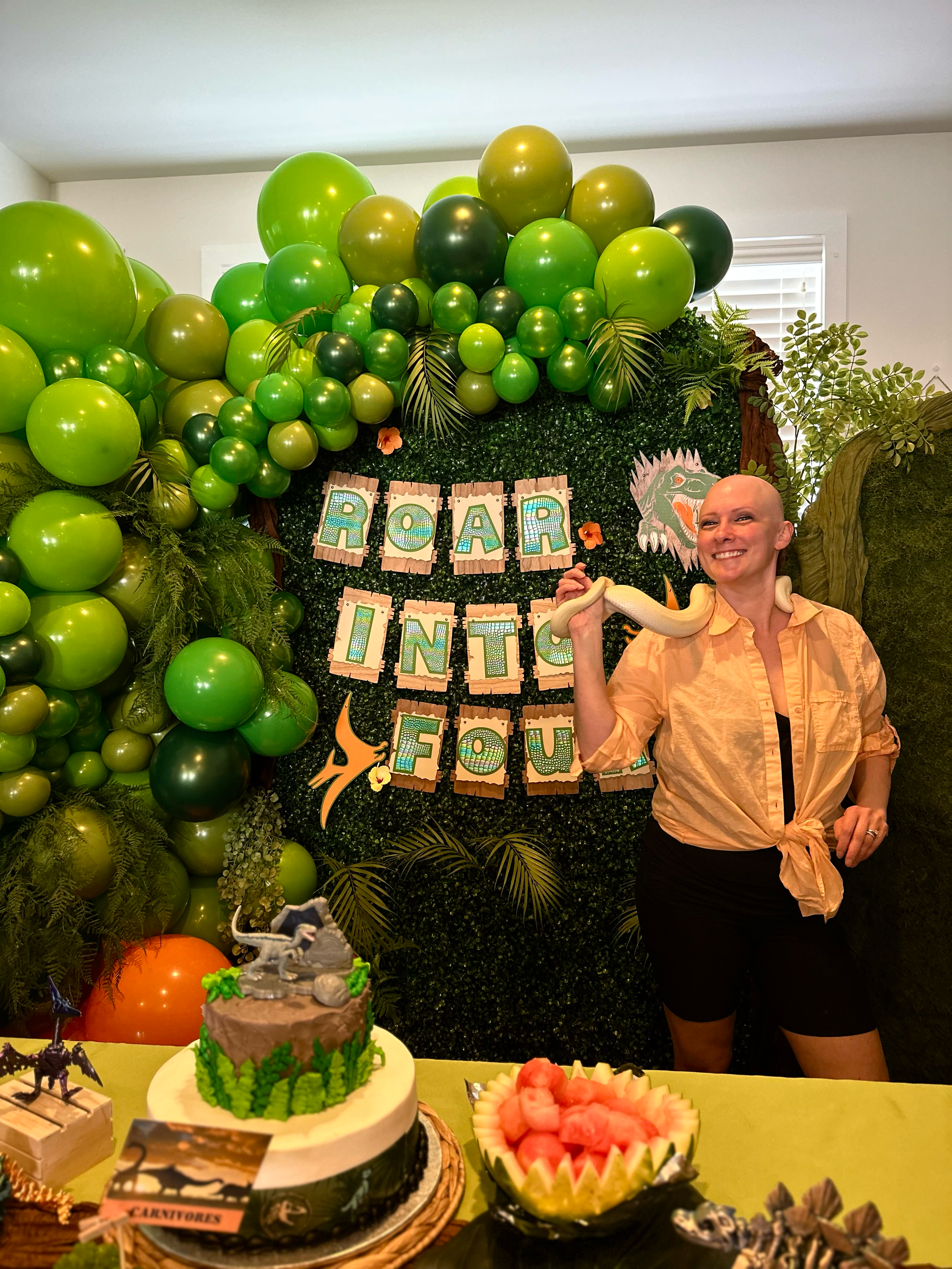 woman holding a snake with a roar into four dinosaur backdrop with green balloons.
