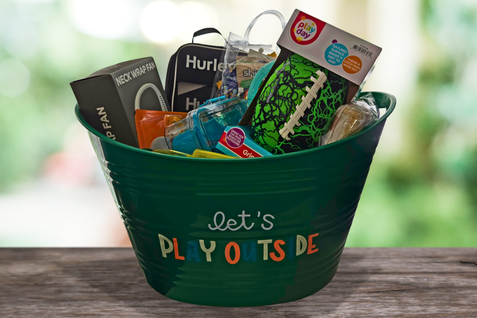 Green bucket that reads Let's Play Outside, filled with outdoor toys, a neck fan, football, and a snackle box.