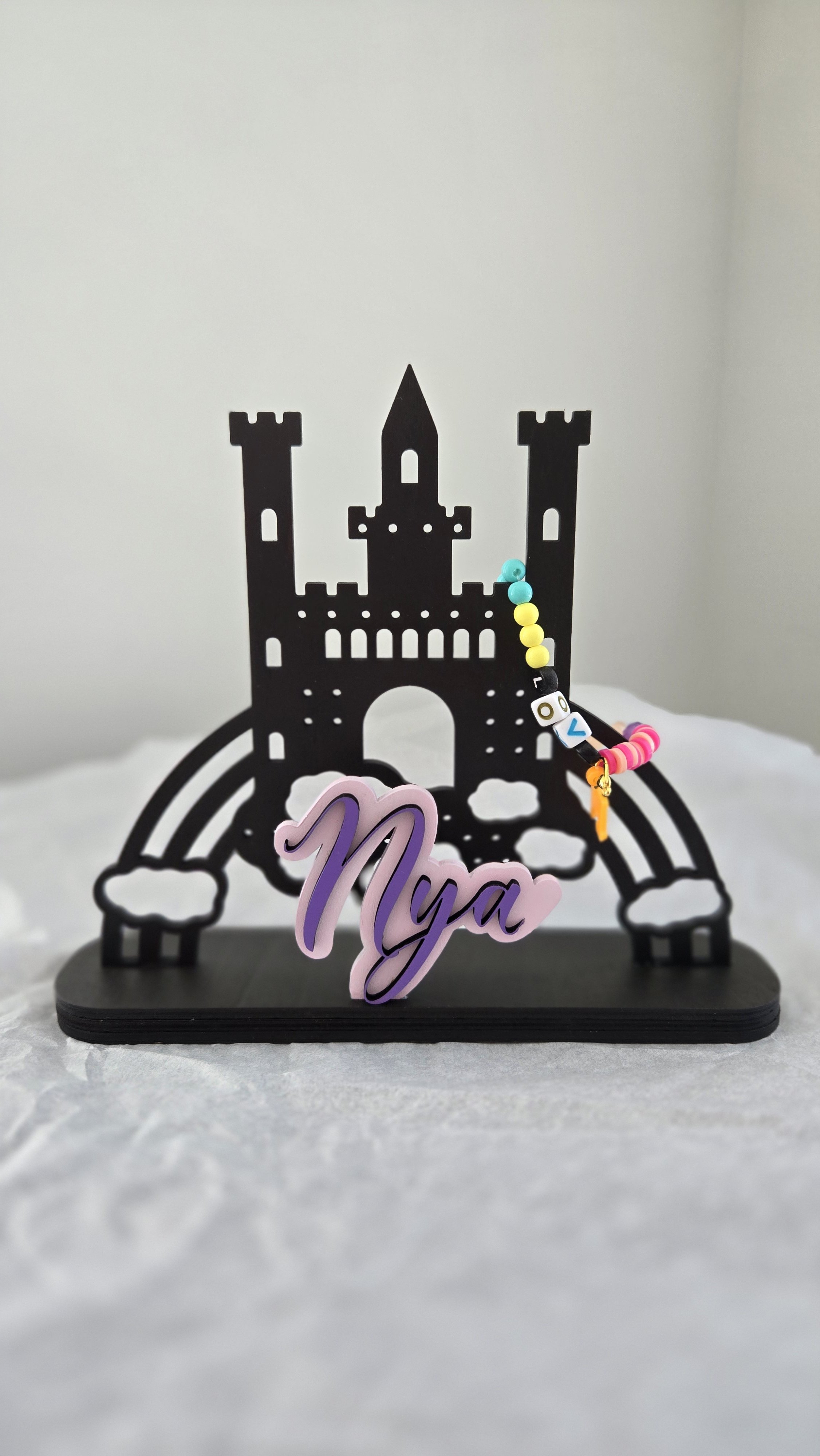 Black castle-shaped jewelry holder with beads attached and a pink decoration with the name 'Nya' on it, placed on a white surface with a plain background.