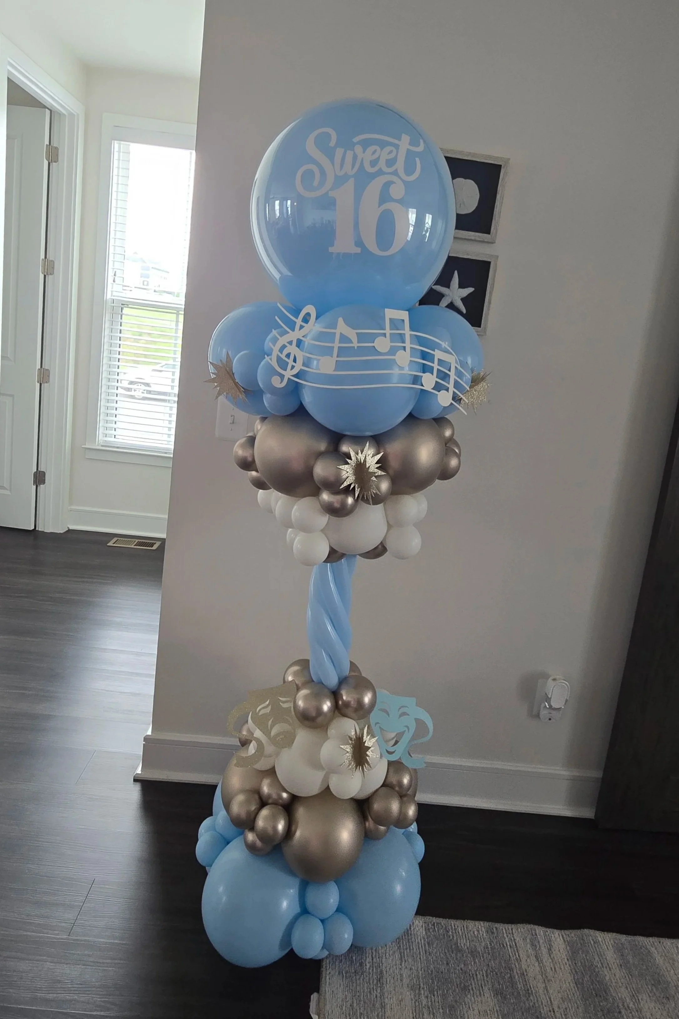 Balloon arrangement celebrating a 16th birthday with blue, white, and silver balloons, musical notes, and a "Sweet 16" sign.