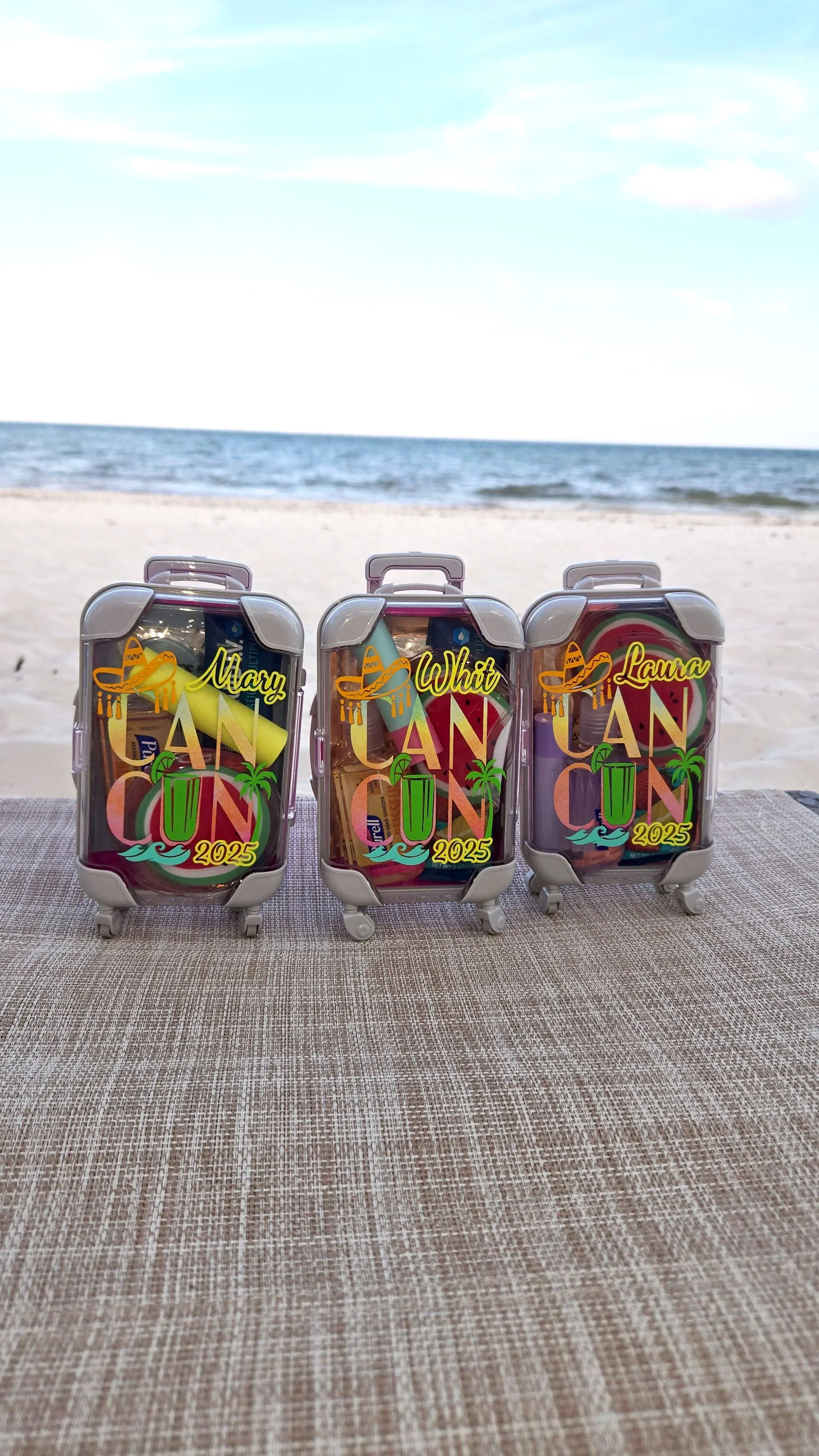 Three mini suitcase favor boxes personalized with names and Cancun 2025, filled with travel essentials, on a beach