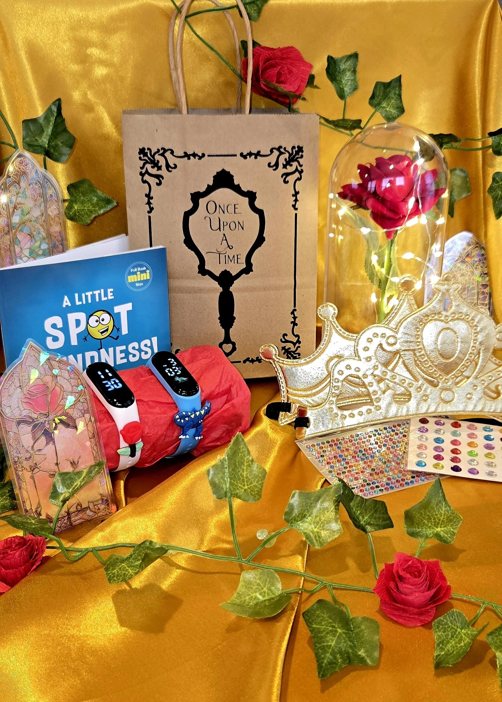 Decorative display with a ribbon, a red rose, a paper bag with a "Once Upon a Time" label, a booklet titled "A Little Spot of Kindness," a plastic dome with a red flower inside, gold decorative items, colorful stickers, and green ivy on a shiny gold fabric background.