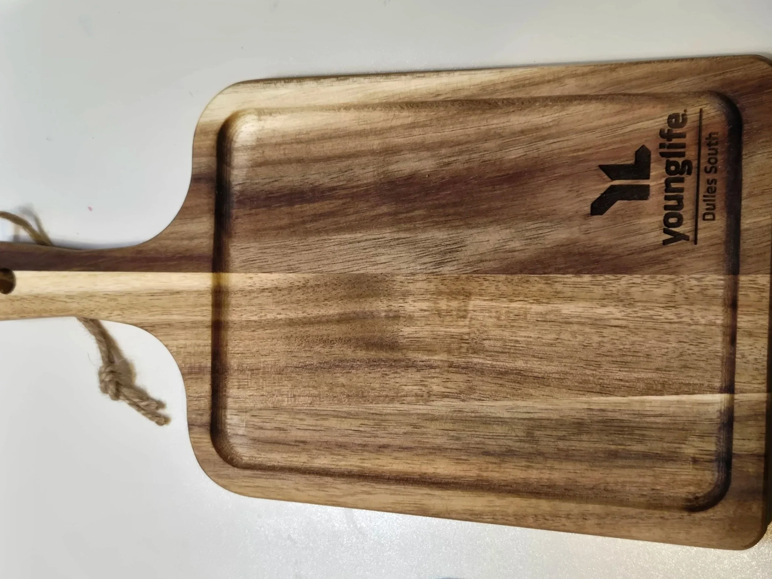 Acacia paddle serving board with a jute rope handle, engraved with the Young Life Dulles South logo