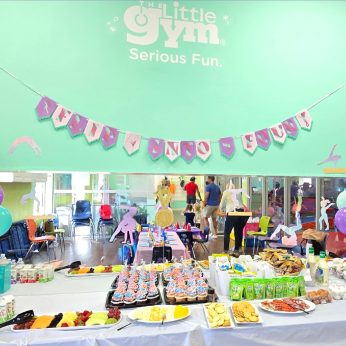 A birthday party at The Little Gym with colorful decorations, a table of snacks and cupcakes, and children and adults in the background. Display of custom made banner, wall embellishments and cupcake toppers.