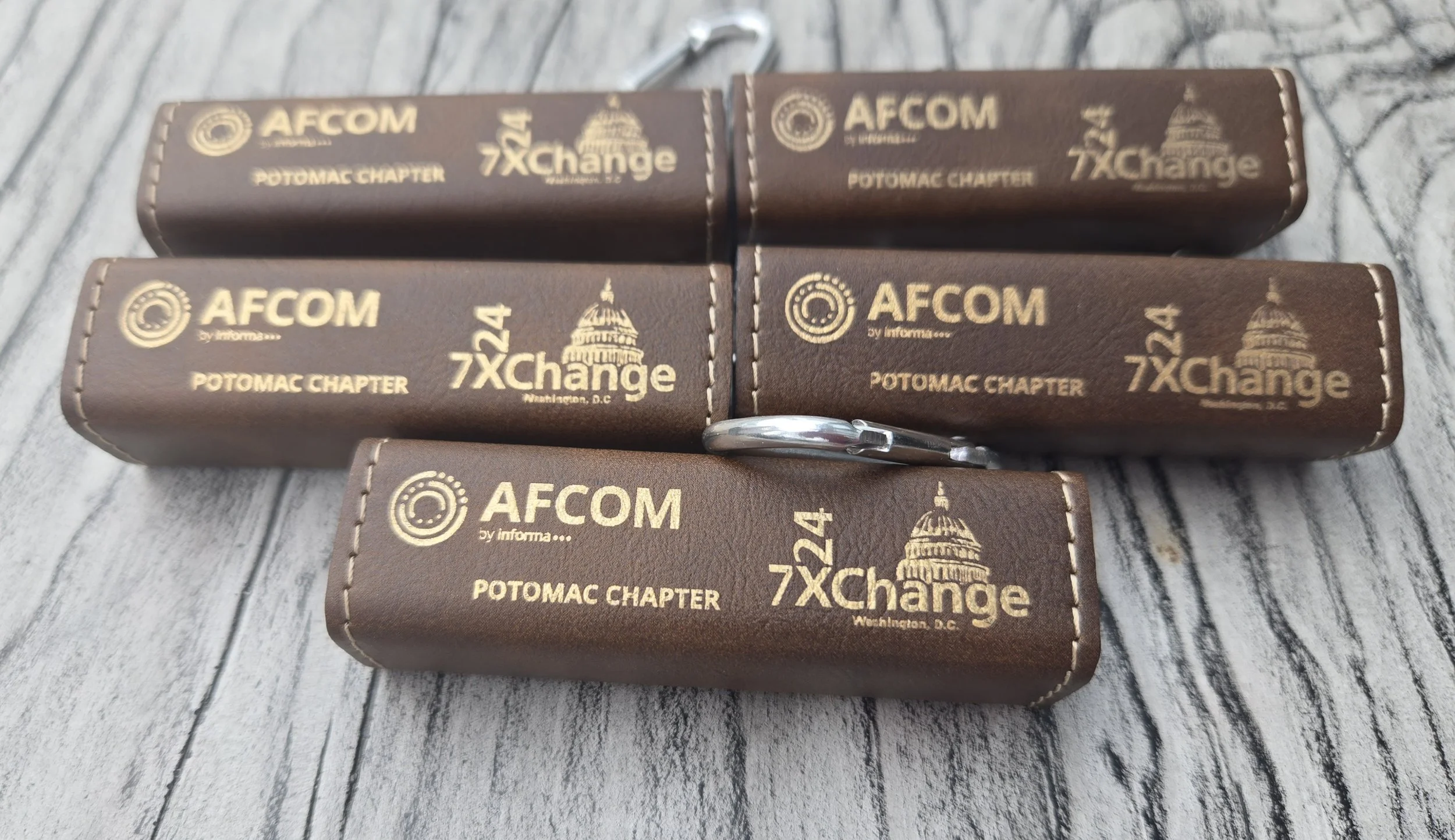 Six brown leather key cases with gold embossed logos and text, placed on a wooden surface. Each case has the logos of AFCOM and 7Xchange, along with additional text indicating they are related to the Potomac Chapter in Washington, D.C.