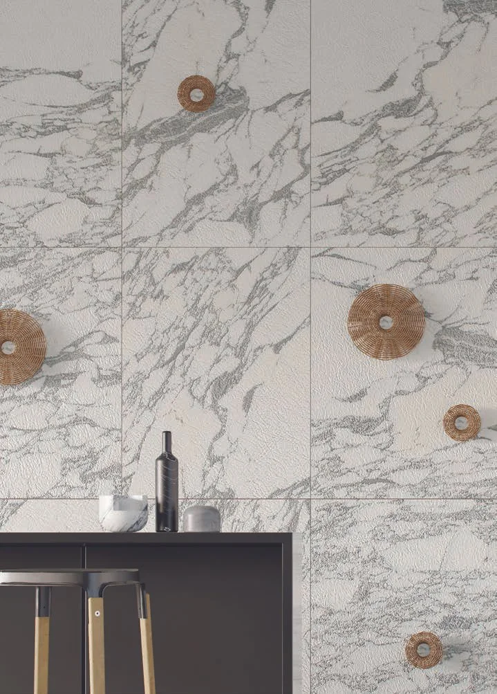 Parete decorata con grandi piastrelle in marmo bianco con venature grigie, decorata con cerchi di vimini di diverse dimensioni. In primo piano, un bancone nero con alcuni oggetti decorativi e un bar stool con seduta in legno.