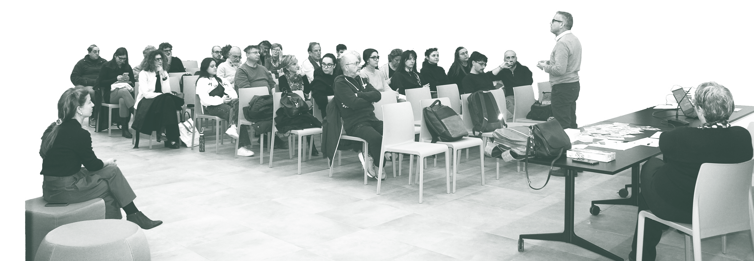 Un relatore sta parlando a un pubblico di adulti seduti in una sala conferenze, alcune persone stanno prendendo appunti o usando il telefono