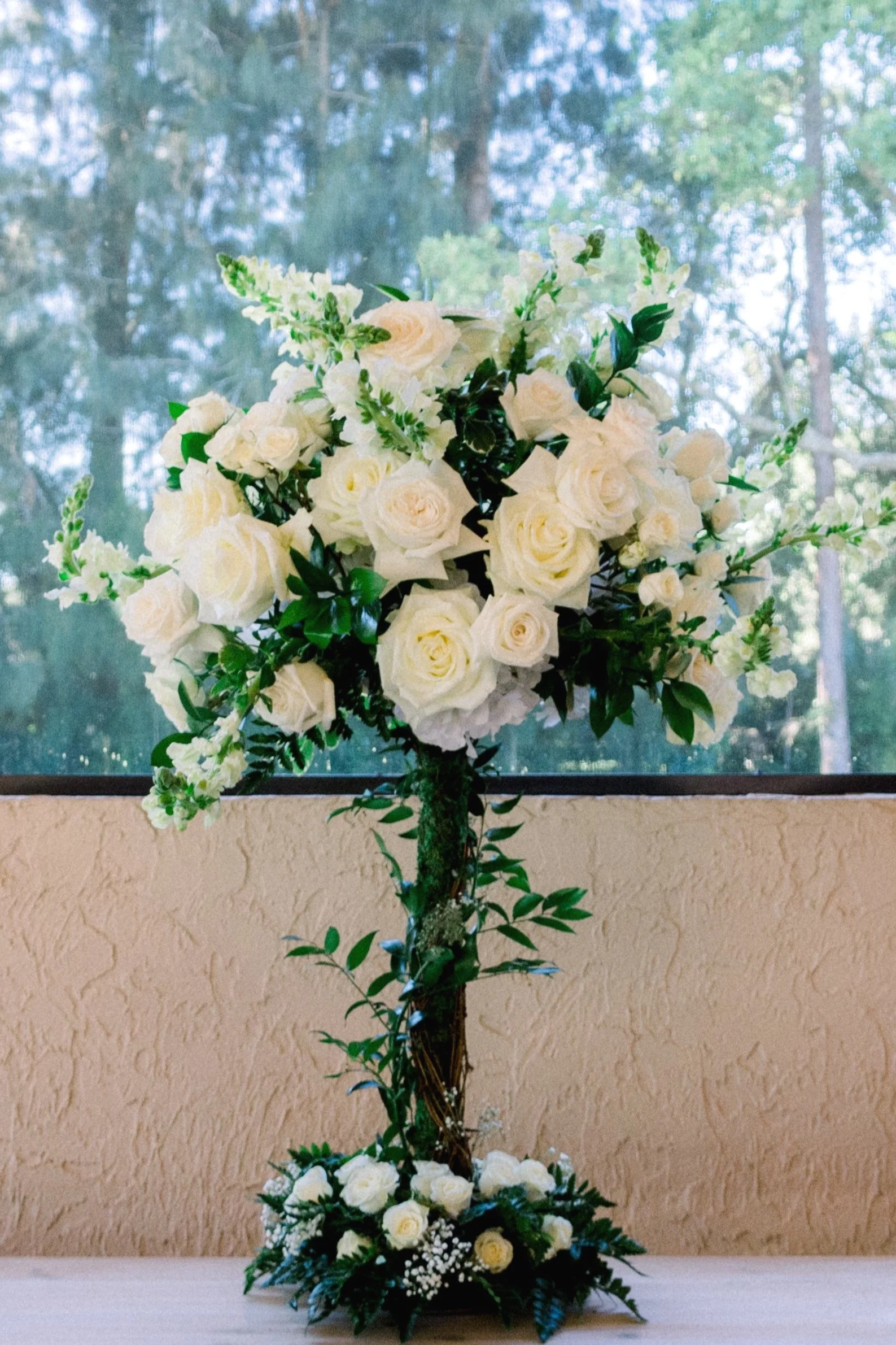 A bouquet display of white roses and greenery on a tall stand against a natural background.