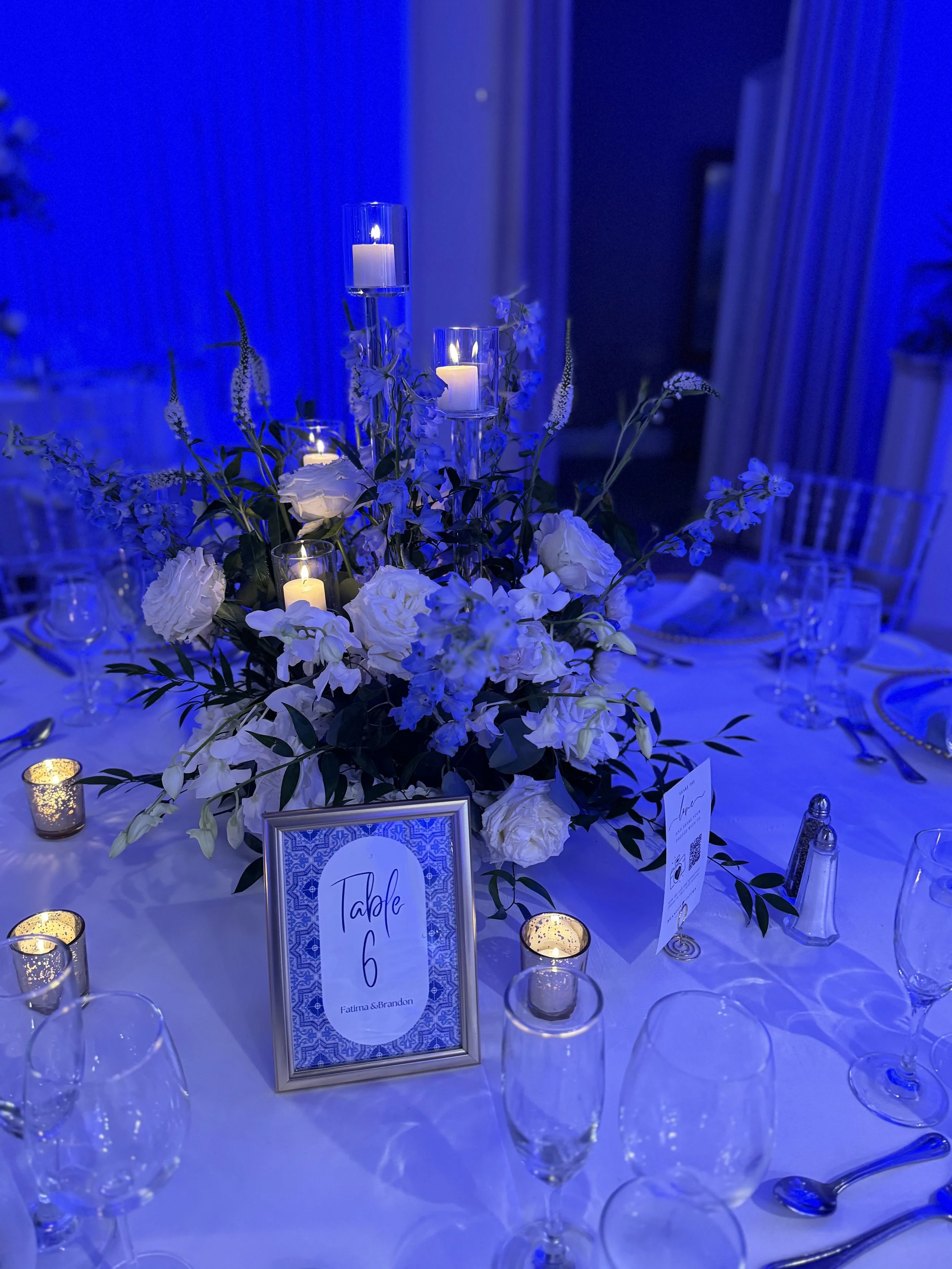 Elegant table setting with a floral centerpiece, candles, and a table number card. The lighting is blue, creating a sophisticated ambiance.