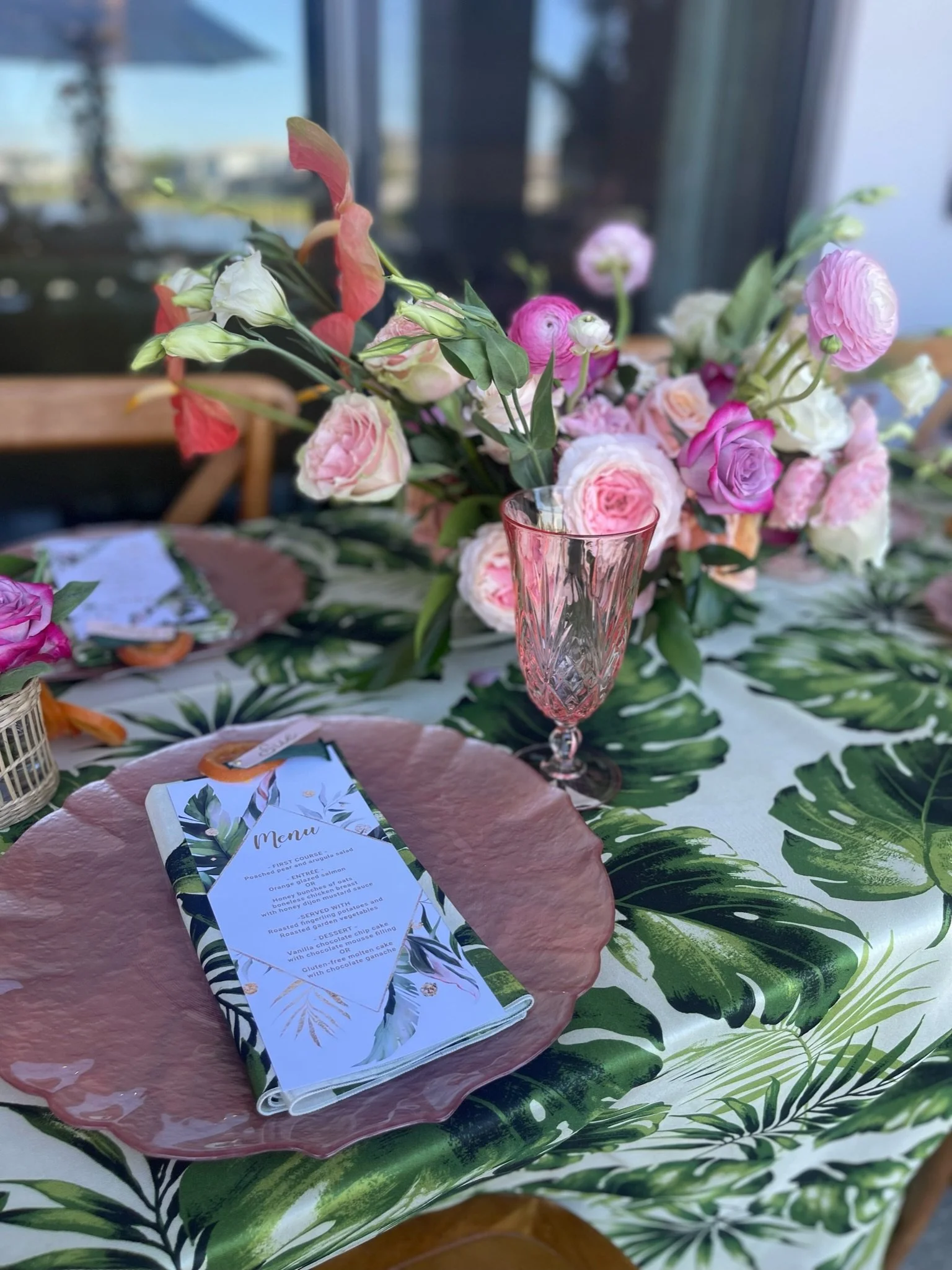 Table setting with tropical-themed decor, pink floral centerpiece, large leaf-patterned tablecloth, pink textured plate, and a menu.