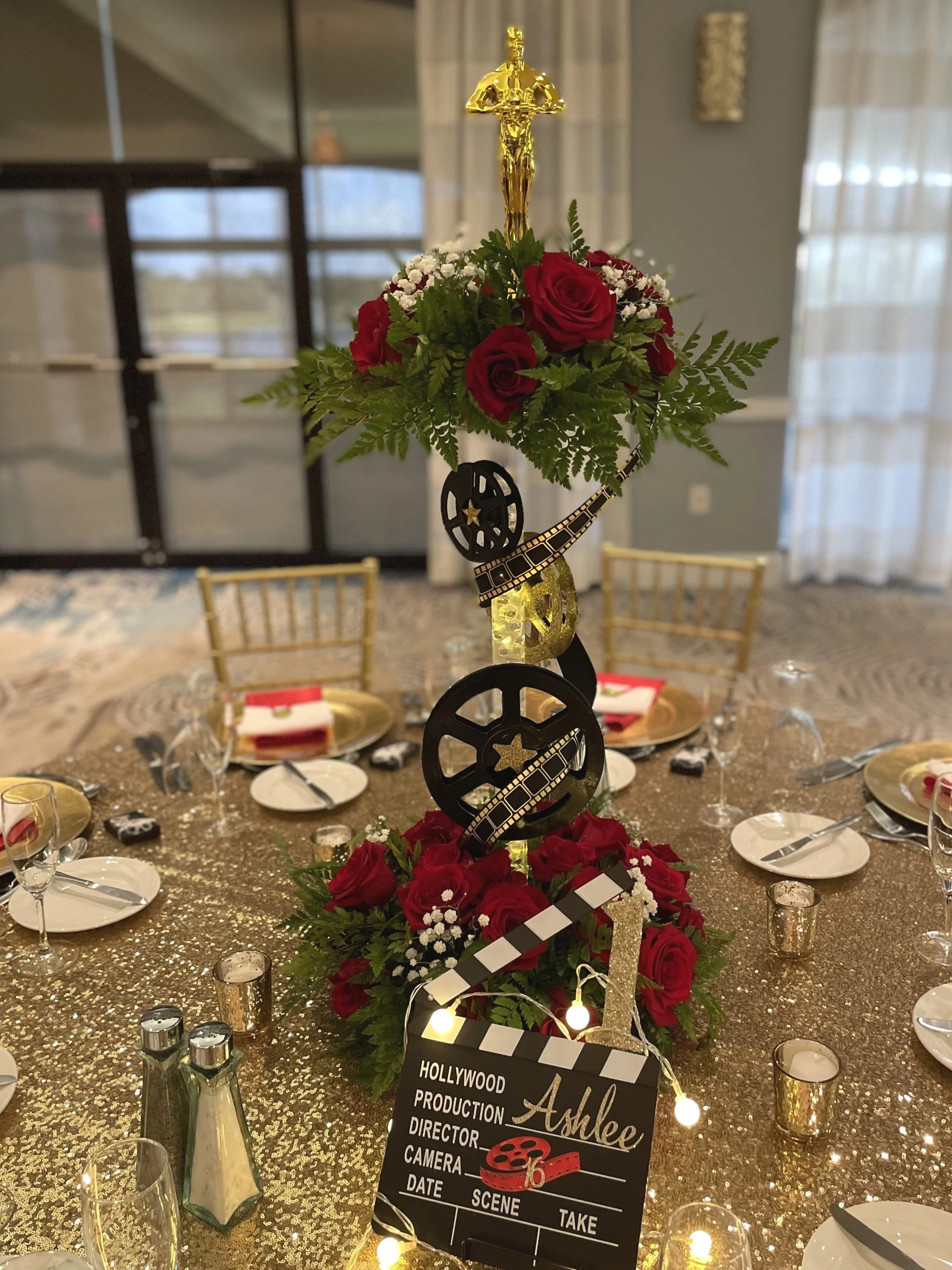 Oscars-themed centerpiece with gold statue, film reel, clapperboard, red roses, and greenery on a glittery table at an event.