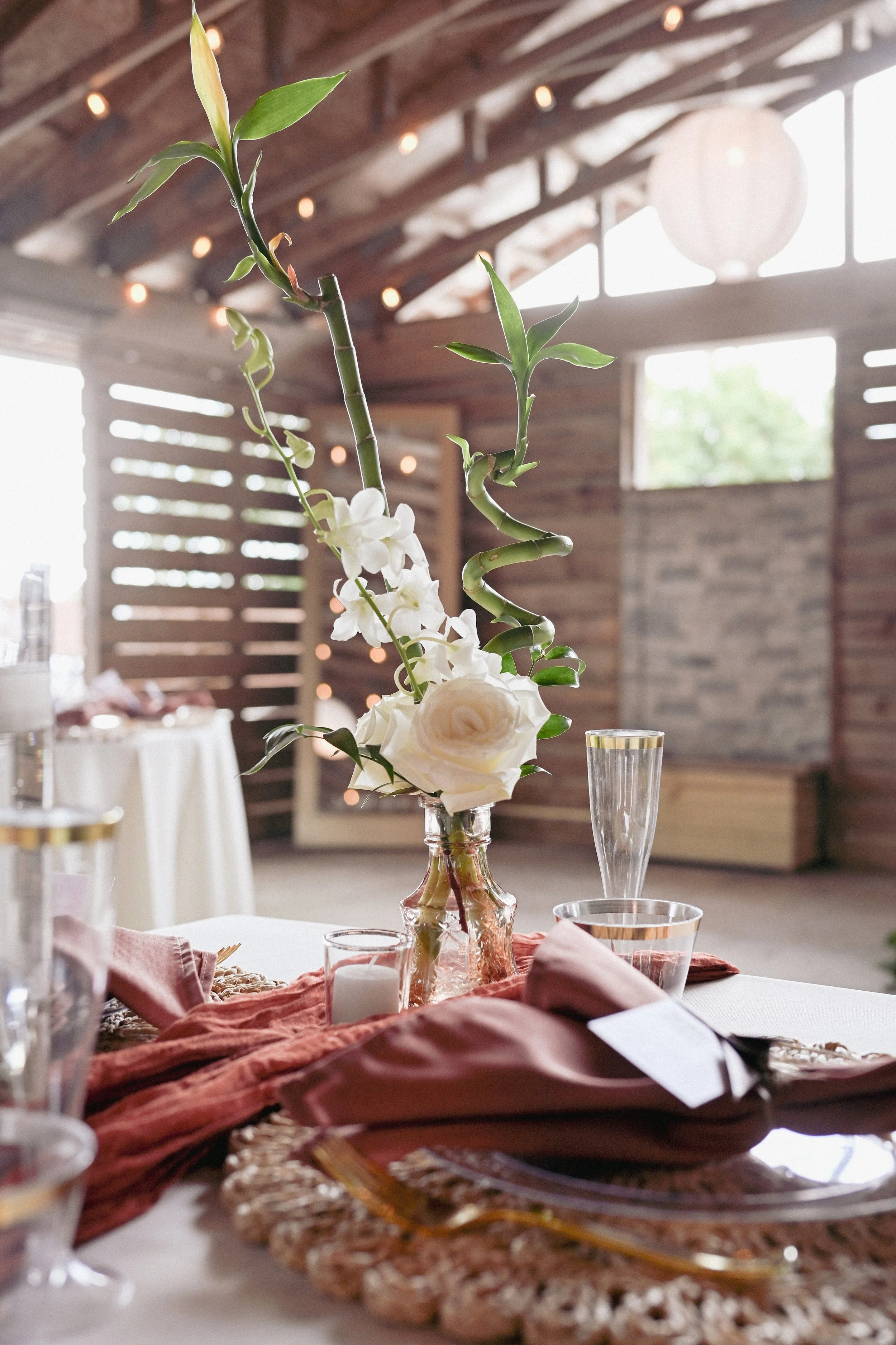 A rustic indoor setting with a table centerpiece featuring a glass vase holding bamboo stalks, white roses, and orchids. The table is decorated with a woven placemat, a folded napkin, glassware, and a candle. Soft lighting and natural wood elements create a cozy atmosphere.
