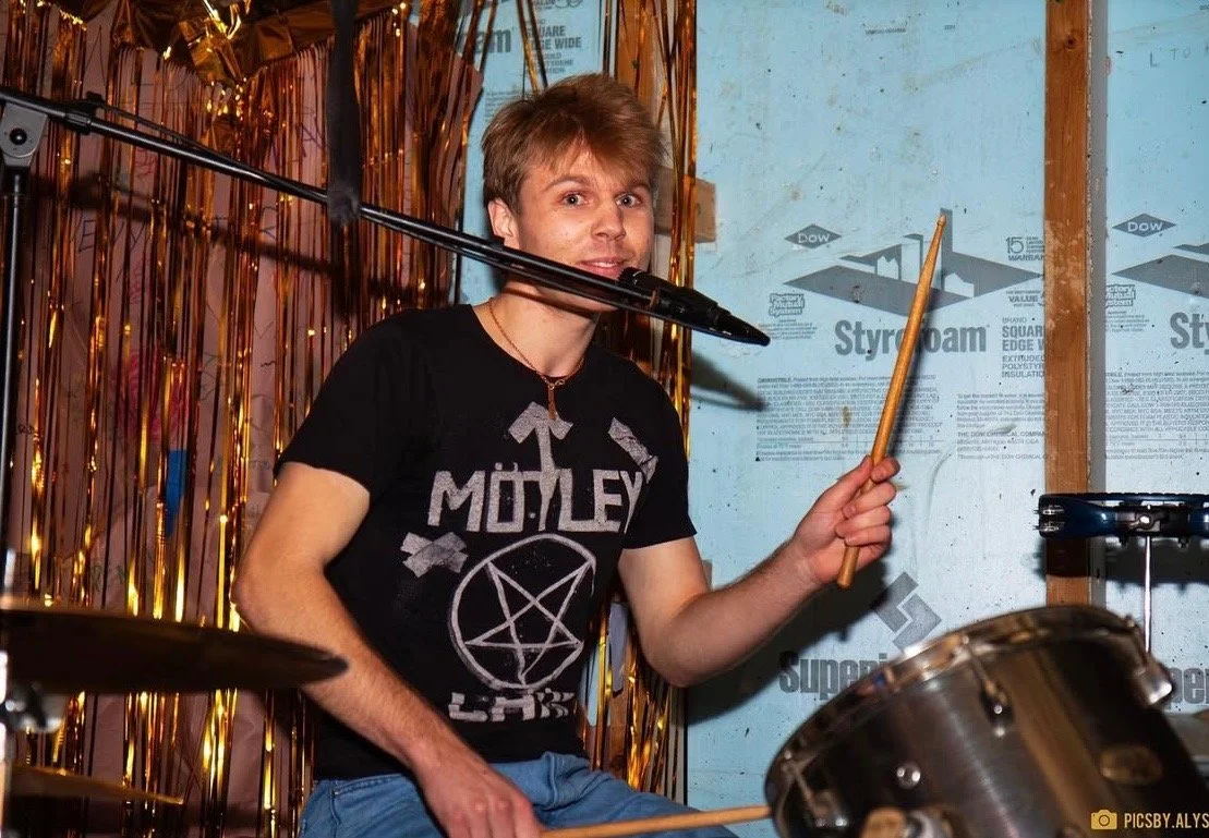 A young person playing drums, wearing a black "Mötley Crüe" t-shirt, holding drumsticks with a microphone positioned in front. The background includes gold streamers and insulation material.