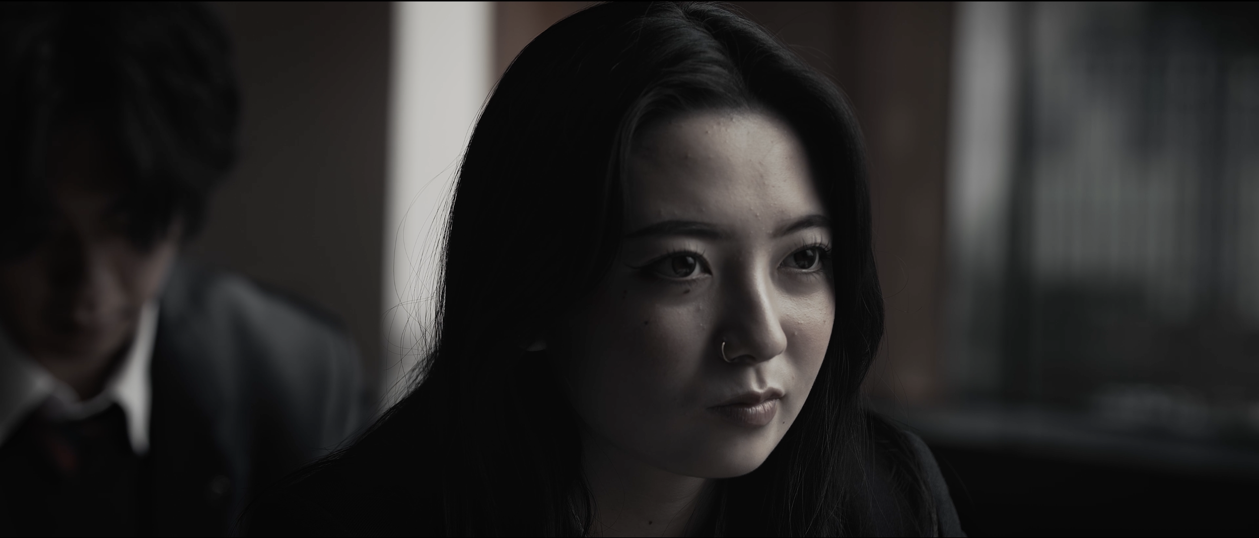Close-up of a woman with long black hair, light skin, wearing a nose ring, looking through a window with a thoughtful expression, in a dimly lit environment.