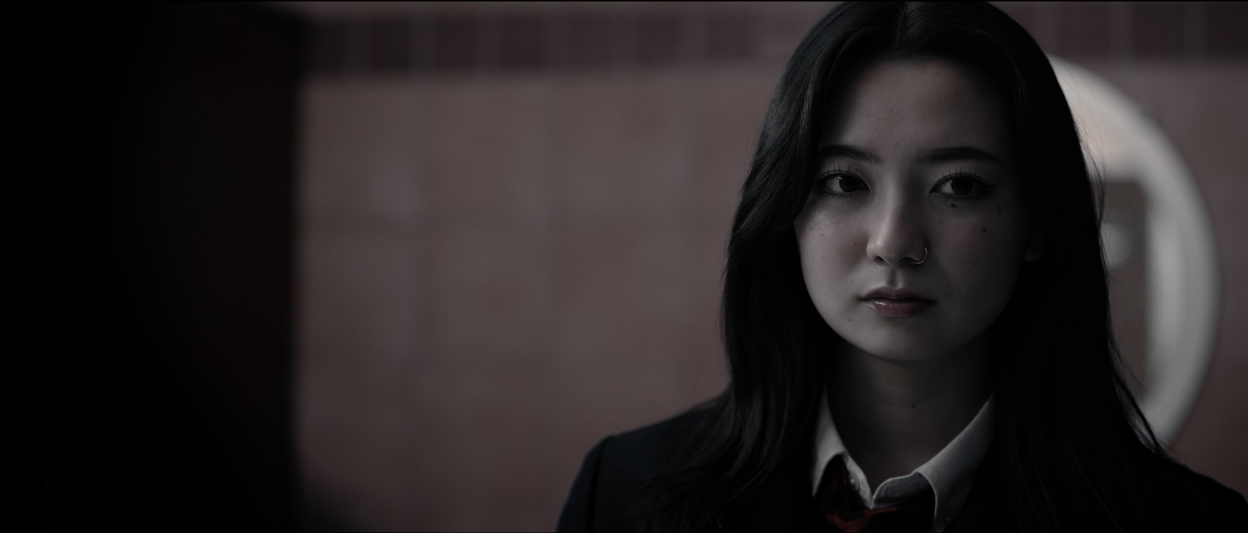 A young woman with dark hair, light skin, and a nose ring looking slightly to the side, wearing a suit and tie, in a dimly lit setting.
