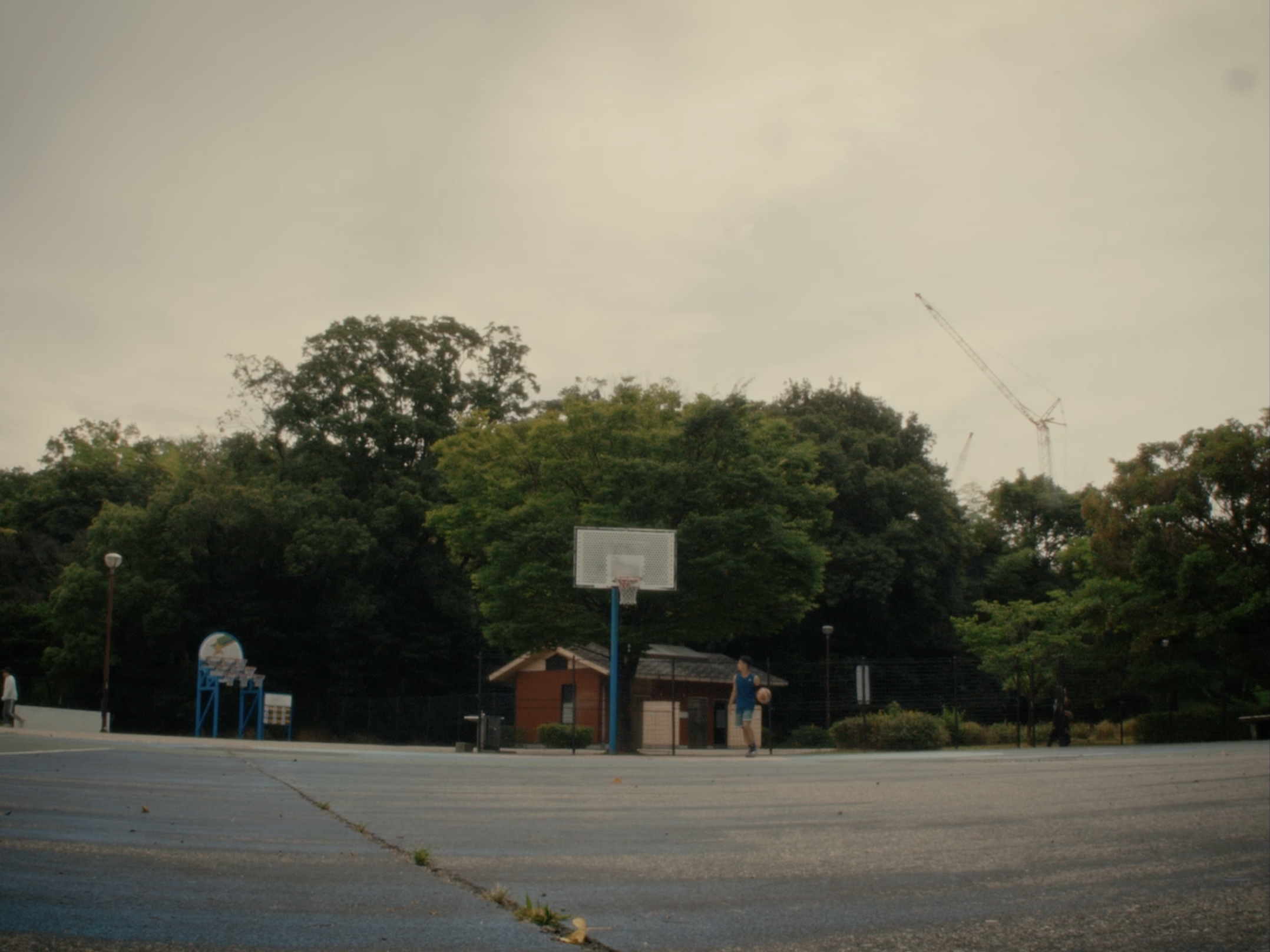 An outdoor basketball court with a hoop in the center, trees and a small building in the background, and a person walking with a soccer ball. The sky is overcast.
