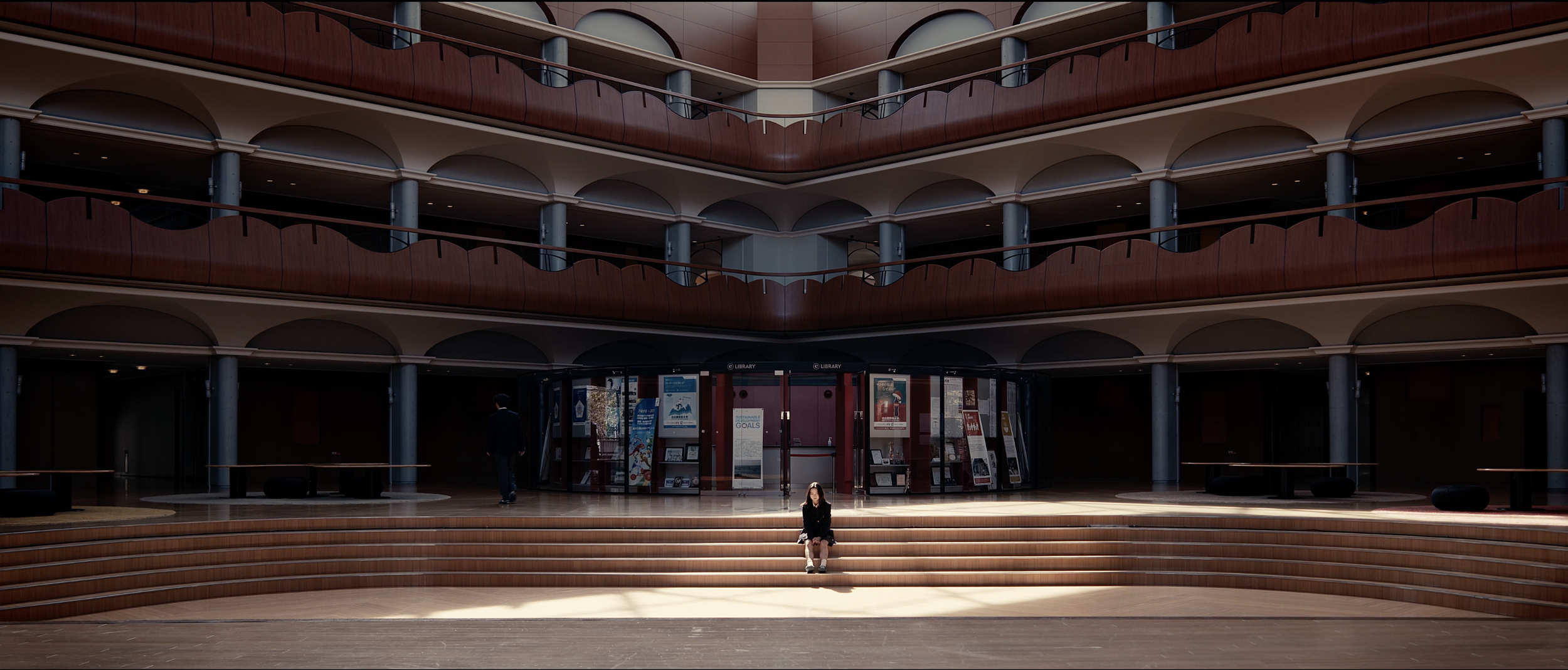 Interior of a modern, multi-story building with a wooden staircase in the center, a woman sitting on the stairs, and a person walking in the background. The building has multiple balconies with wooden railings and arched openings.