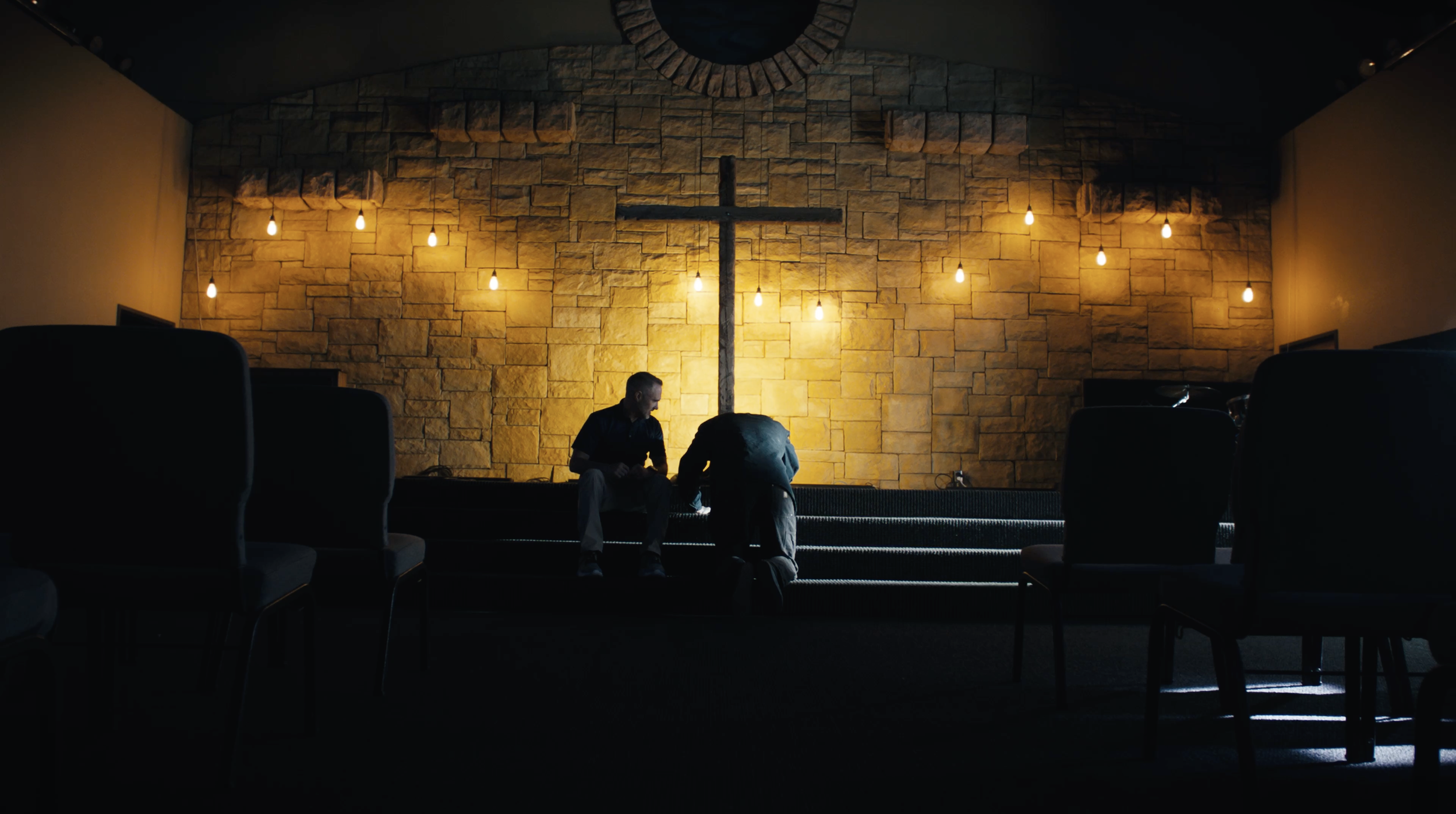 Two individuals sitting on church steps in front of a large cross on a stone wall, with warm hanging lights above, inside a church or chapel.