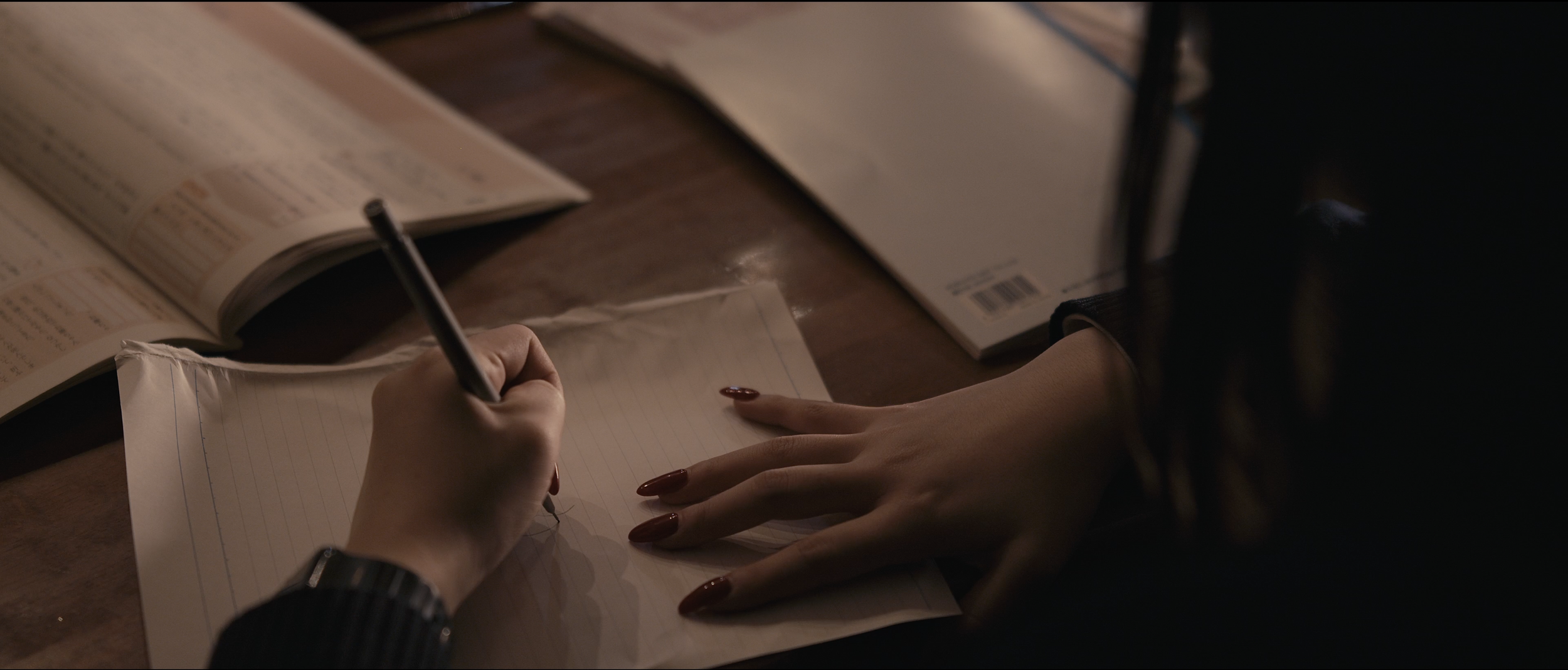 A person writing in a notebook on a wooden table, with open books and papers nearby.