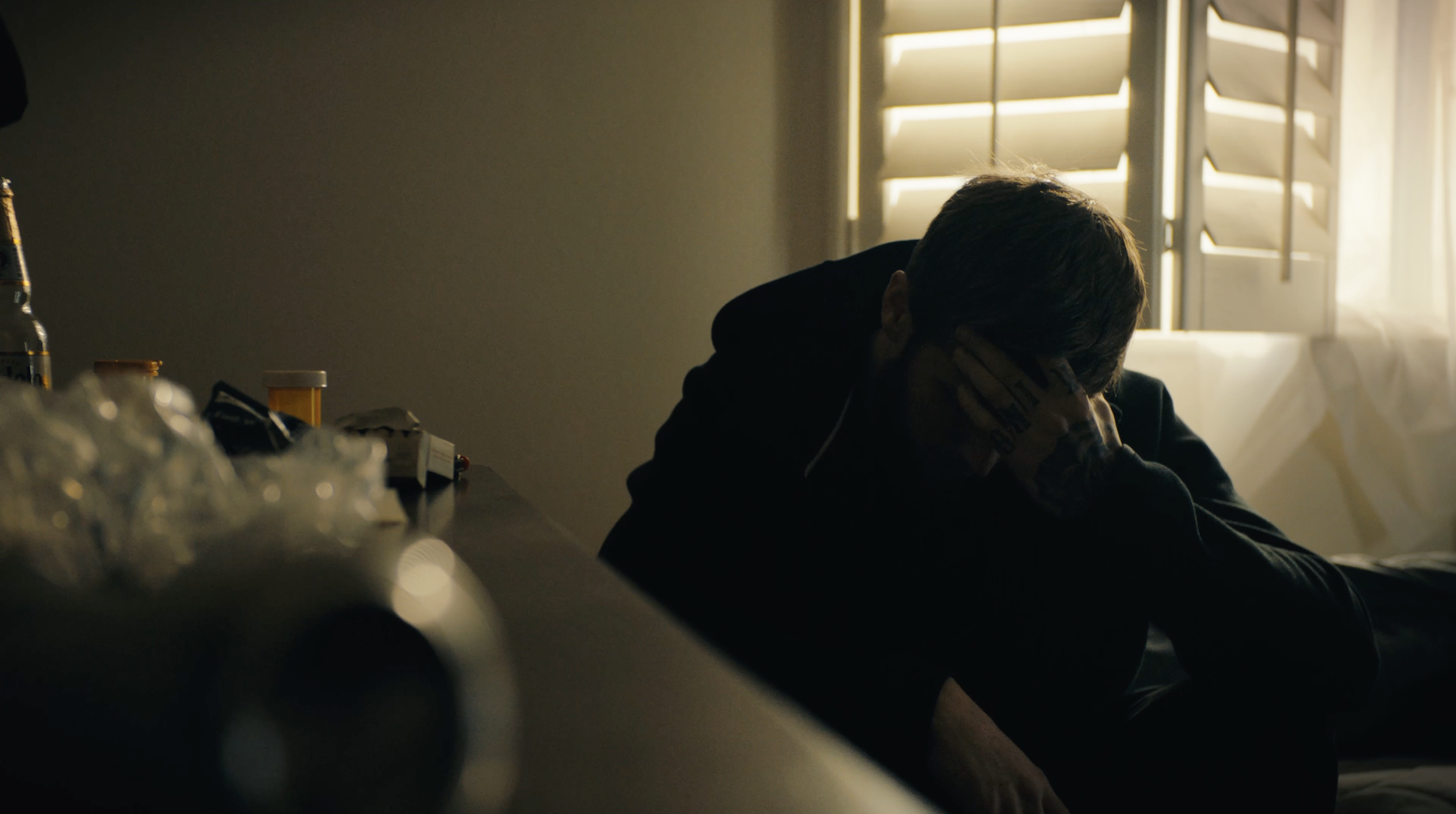 A person with short hair, wearing a dark hoodie, sitting in a dimly lit room with sunlight coming through partially opened shutters, resting their head on their hand.