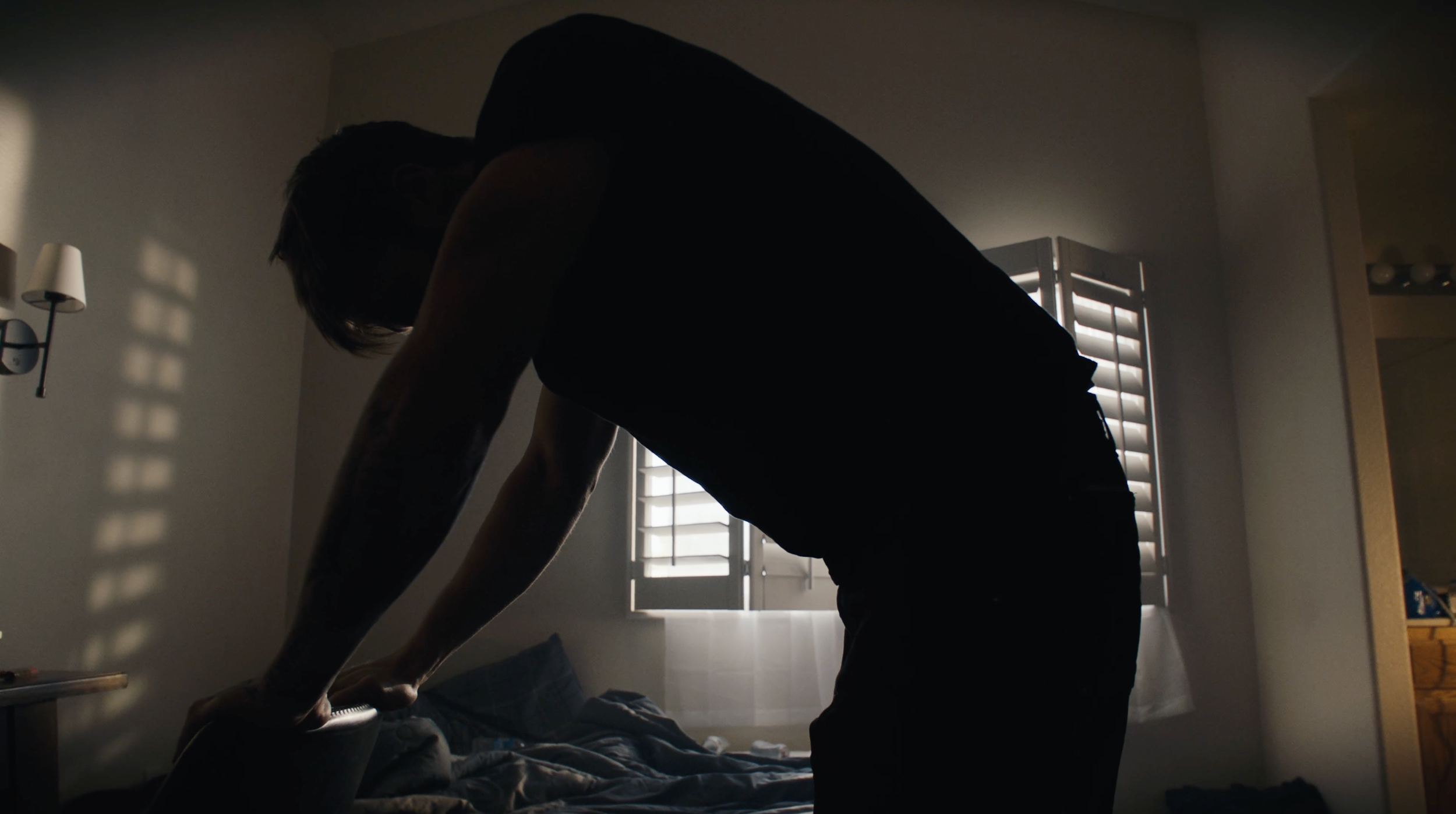Silhouette of a person leaning over a bed in a dimly lit bedroom with sunlight filtering through window shutters.