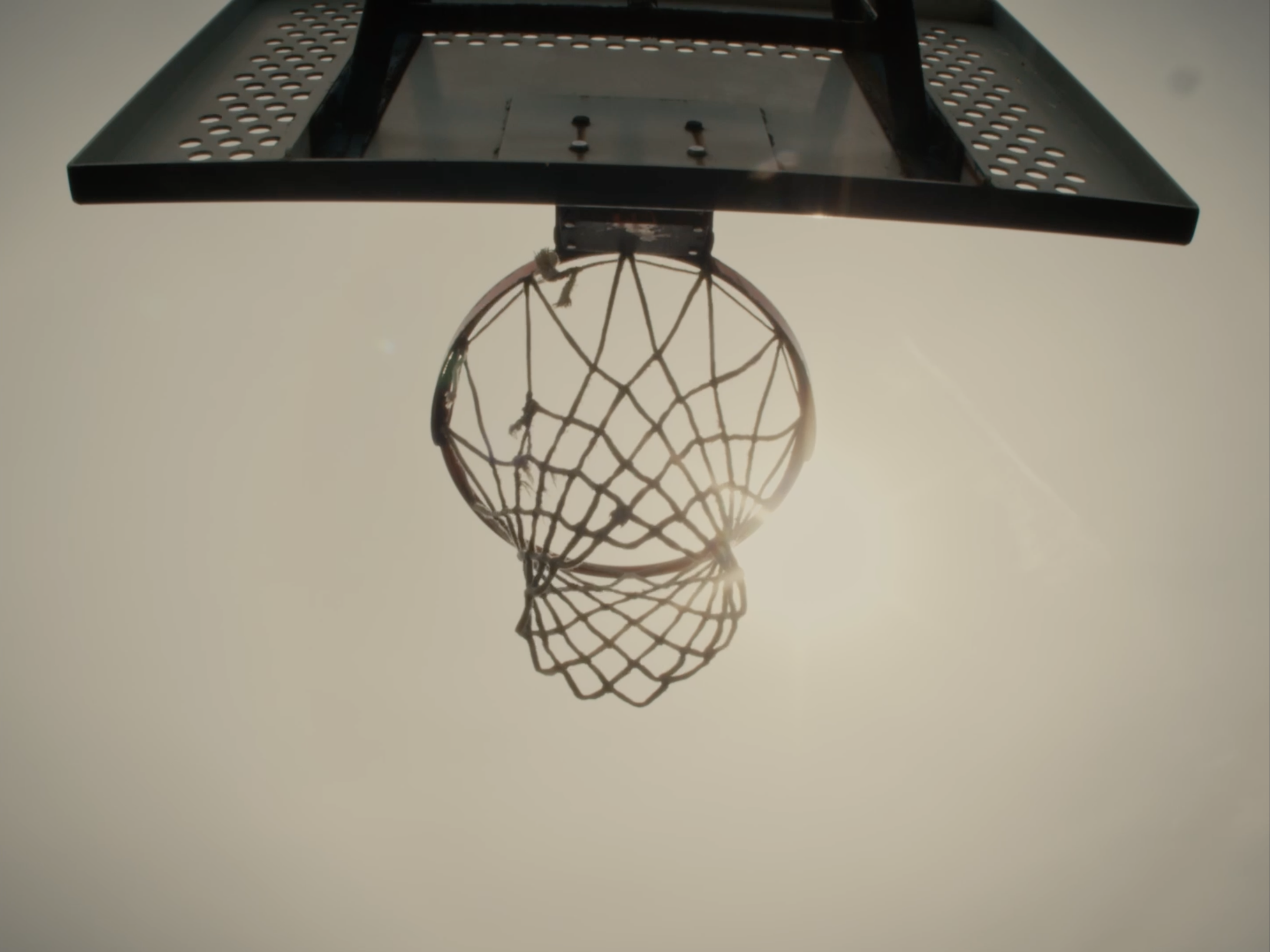A basketball hoop and net from below, with sunlight shining behind the net against a light sky.