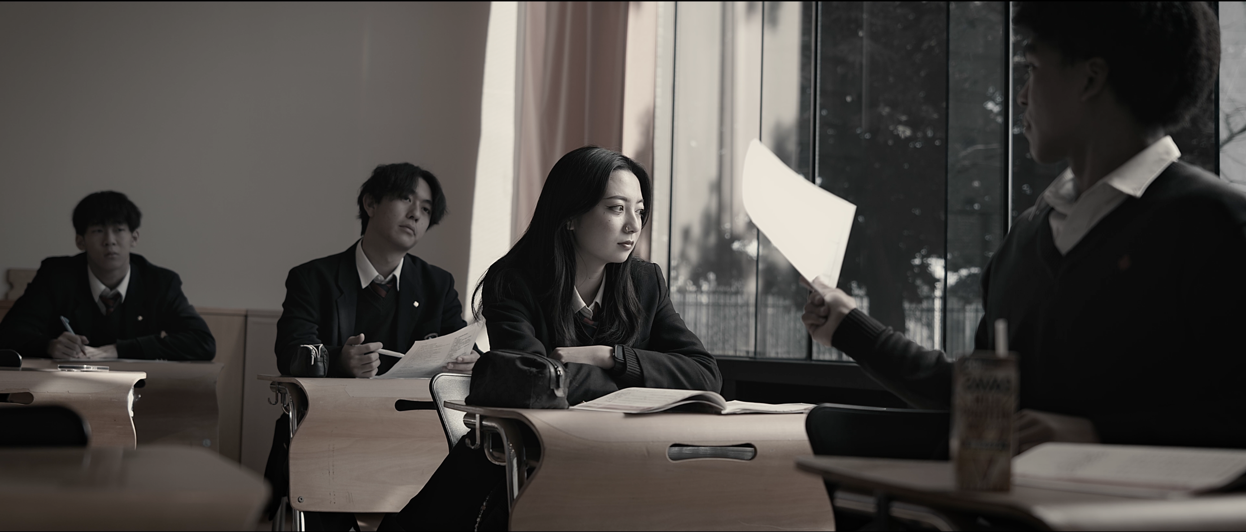 Students in a classroom, one girl attentively listening to a classmate holding a paper, with other students taking notes, natural light coming through large windows.