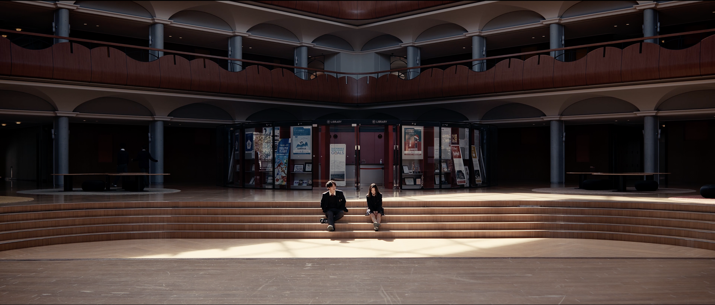 Interior of a modern library with wooden architecture, two students sitting on steps, and posters near the entrance.