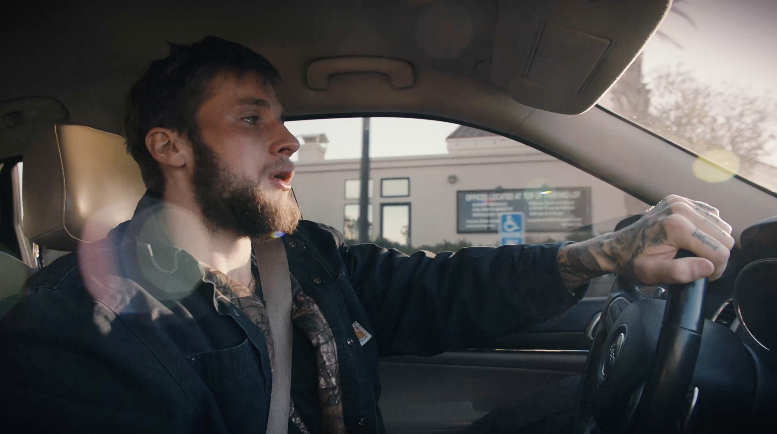 A man with a beard and tattoos, driving a vehicle, wearing a seatbelt, with sunlight coming through the window.