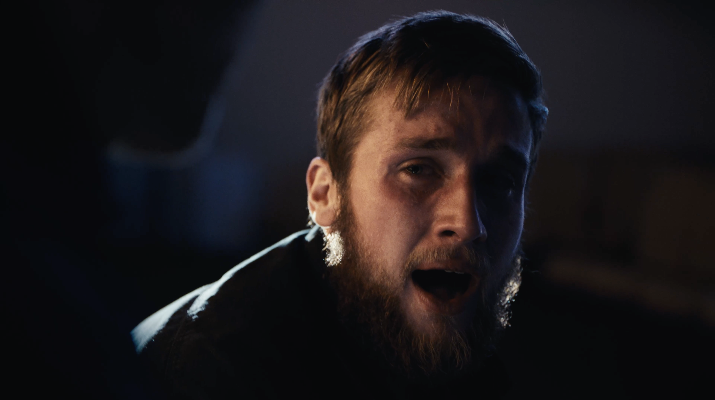 A man with a beard and brown hair has his mouth open, appearing to speak or shout, with a dark background.