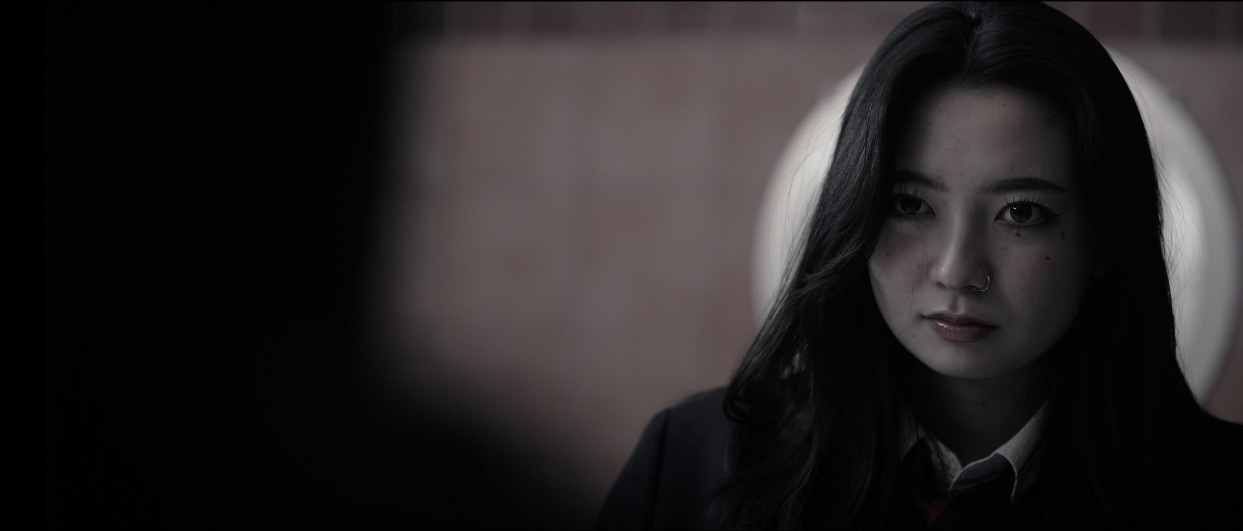 A young woman with long black hair and a nose ring looking at herself in a mirror with a serious expression, in a dimly lit room.