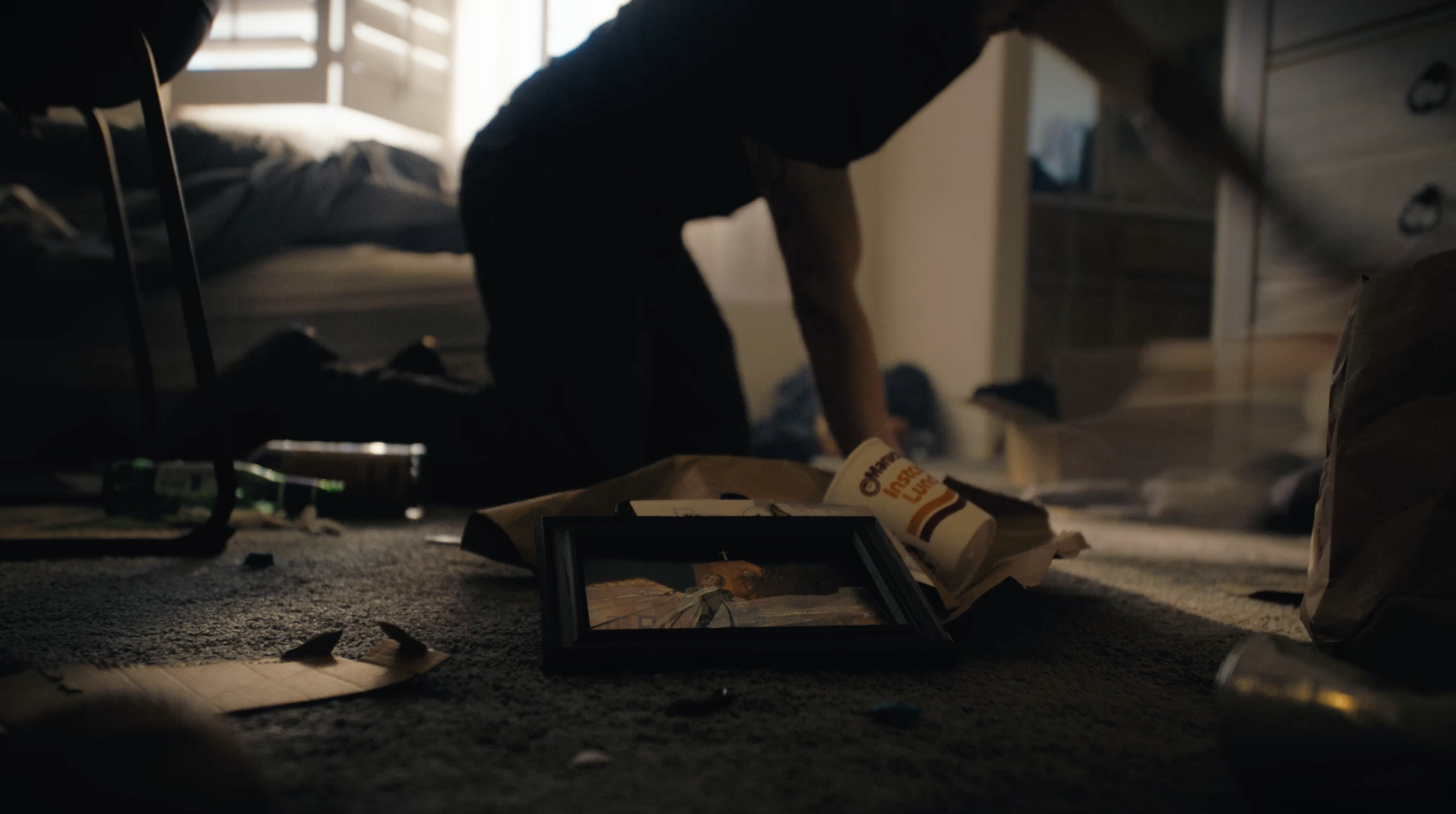 A messy bedroom with a person kneeling on the floor. Items including a framed picture, a McDonald's cup, and scattered trash are visible. Sunlight streams through a window, illuminating the clutter.