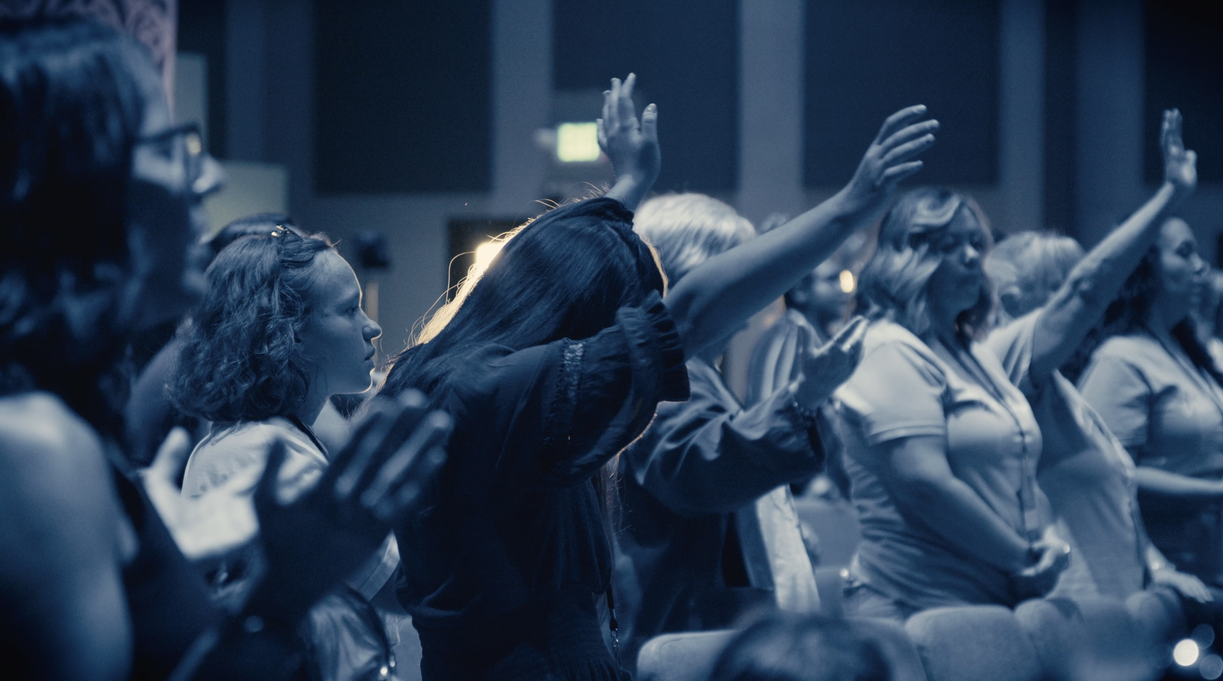 Group of women in an audience raising their hands during an event in a dark room with stage lighting.