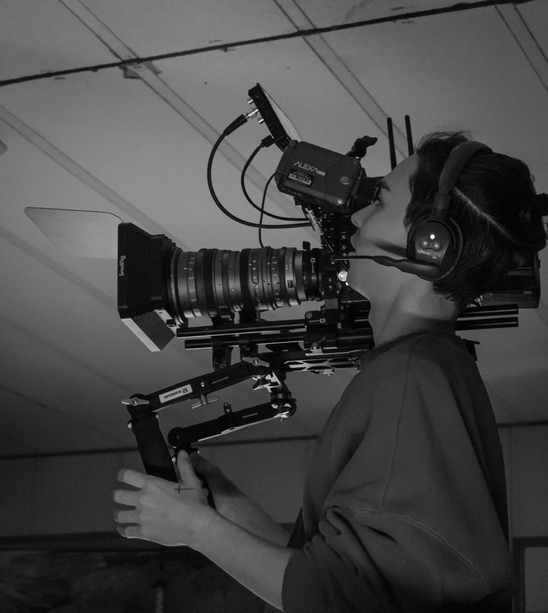 A person wearing headphones operating a professional camera rig with a large lens, mounted on a stabilizer. The scene is in black and white.