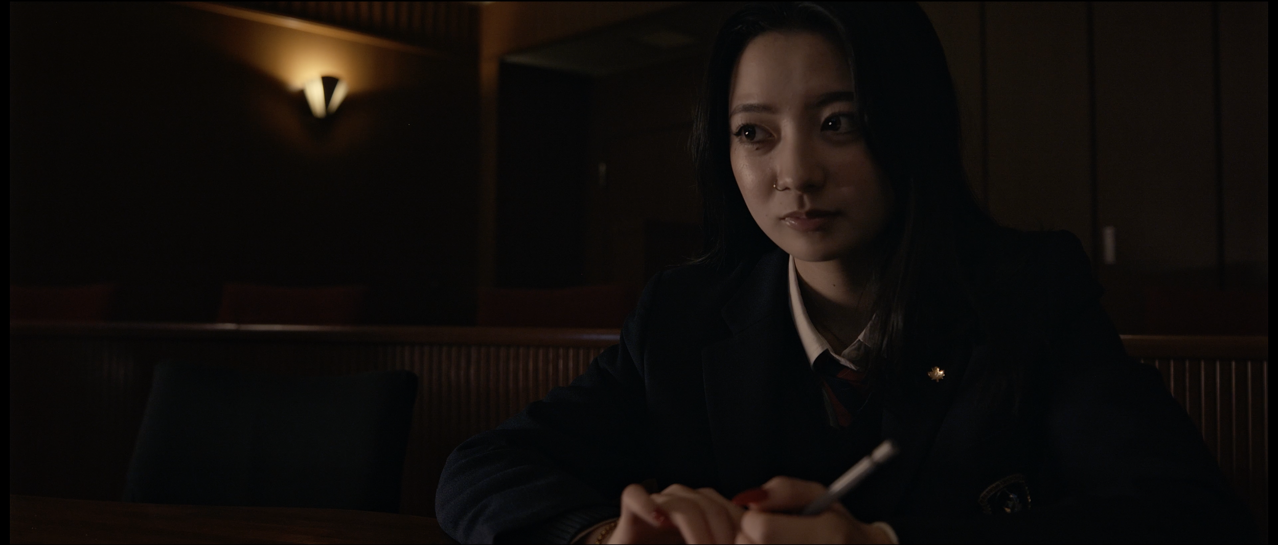 A young woman with dark hair and a nose piercing sitting at a wooden table in a dimly lit room, holding a pen, wearing a dark blazer and a white shirt.