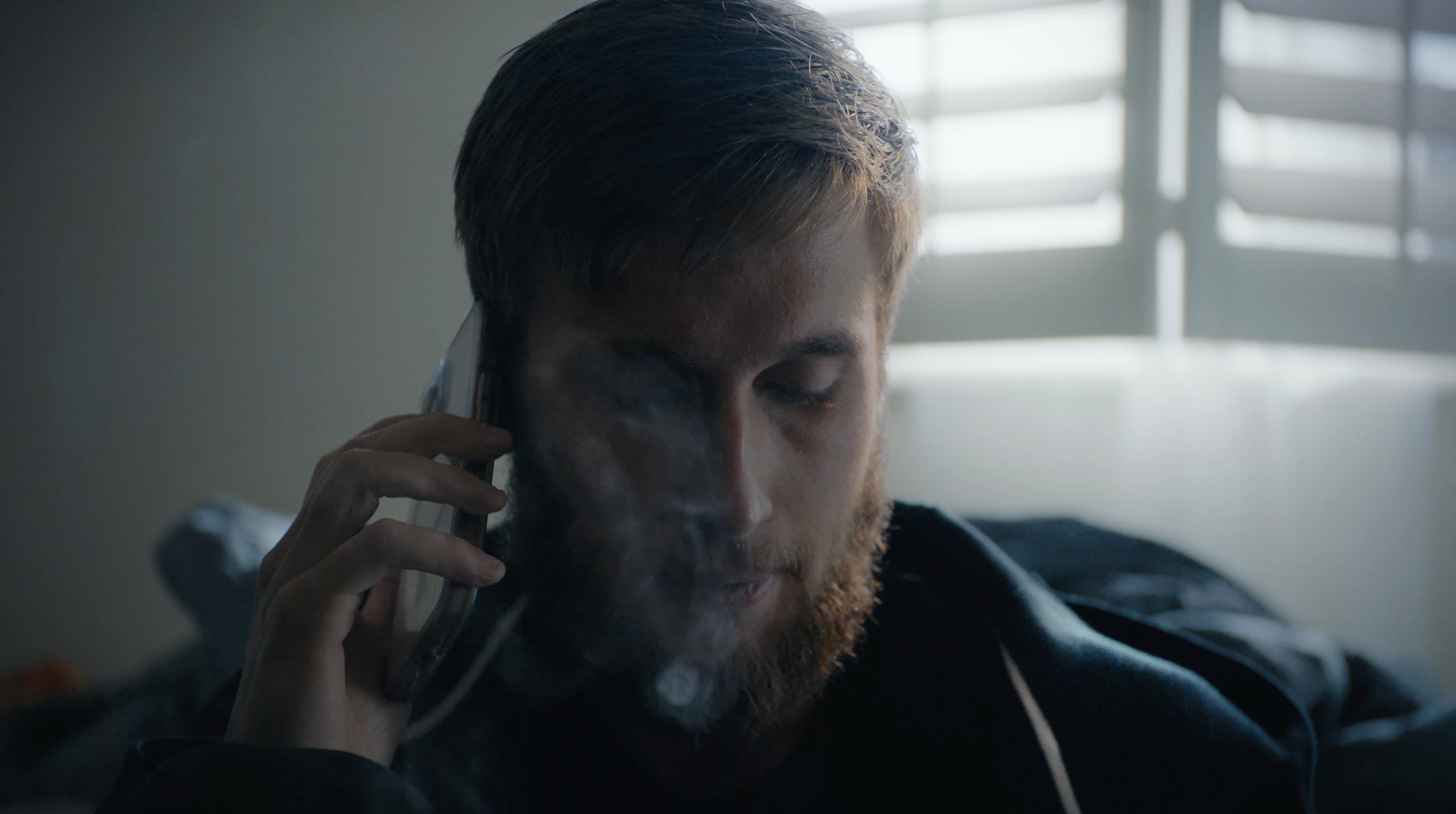 A man with a beard and closed eyes talking on a telephone, with visible smoke near his face, in a room with a window in the background.